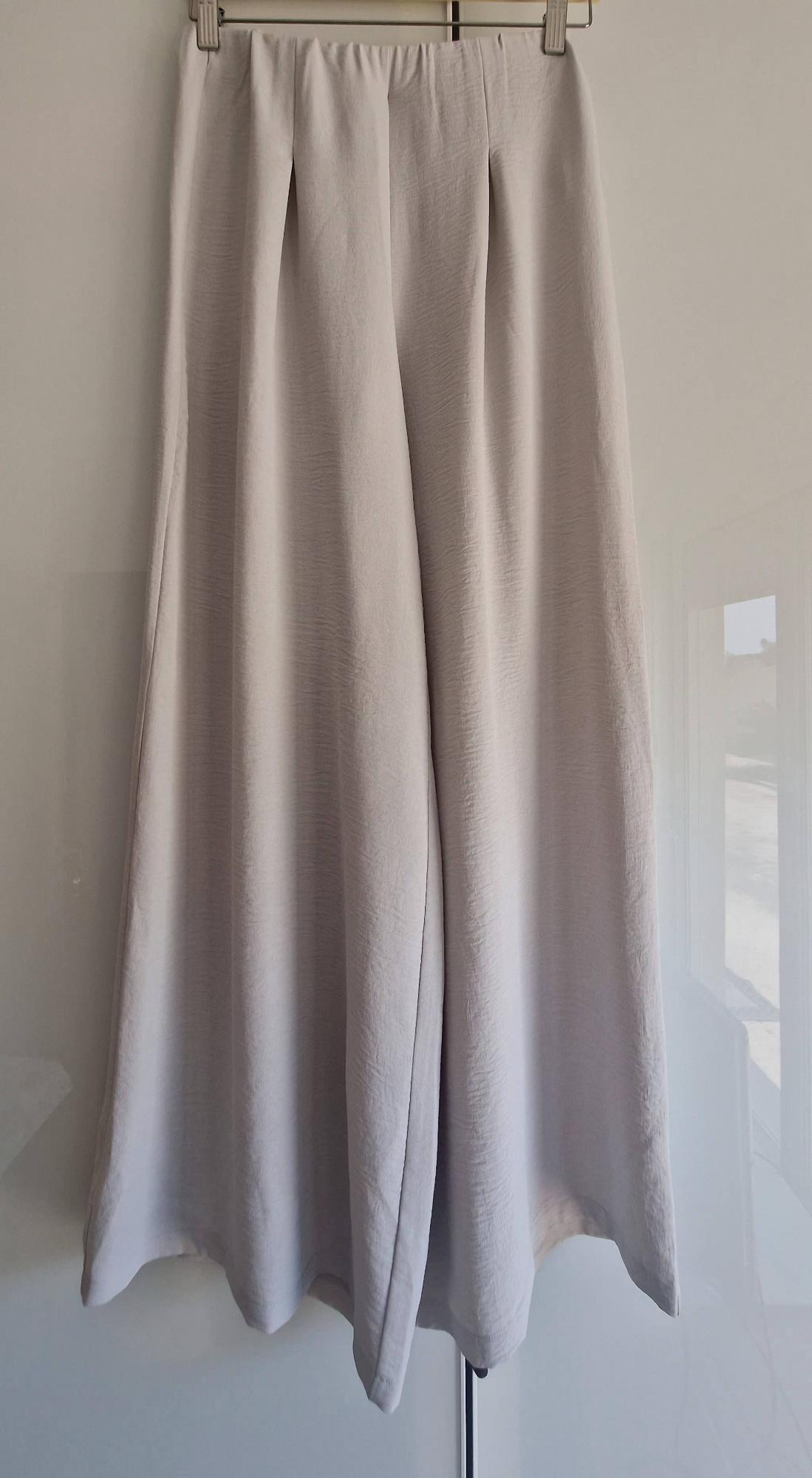 Flowy wide-leg trousers in light beige with pleats, hanging on a hanger above a glossy white surface.