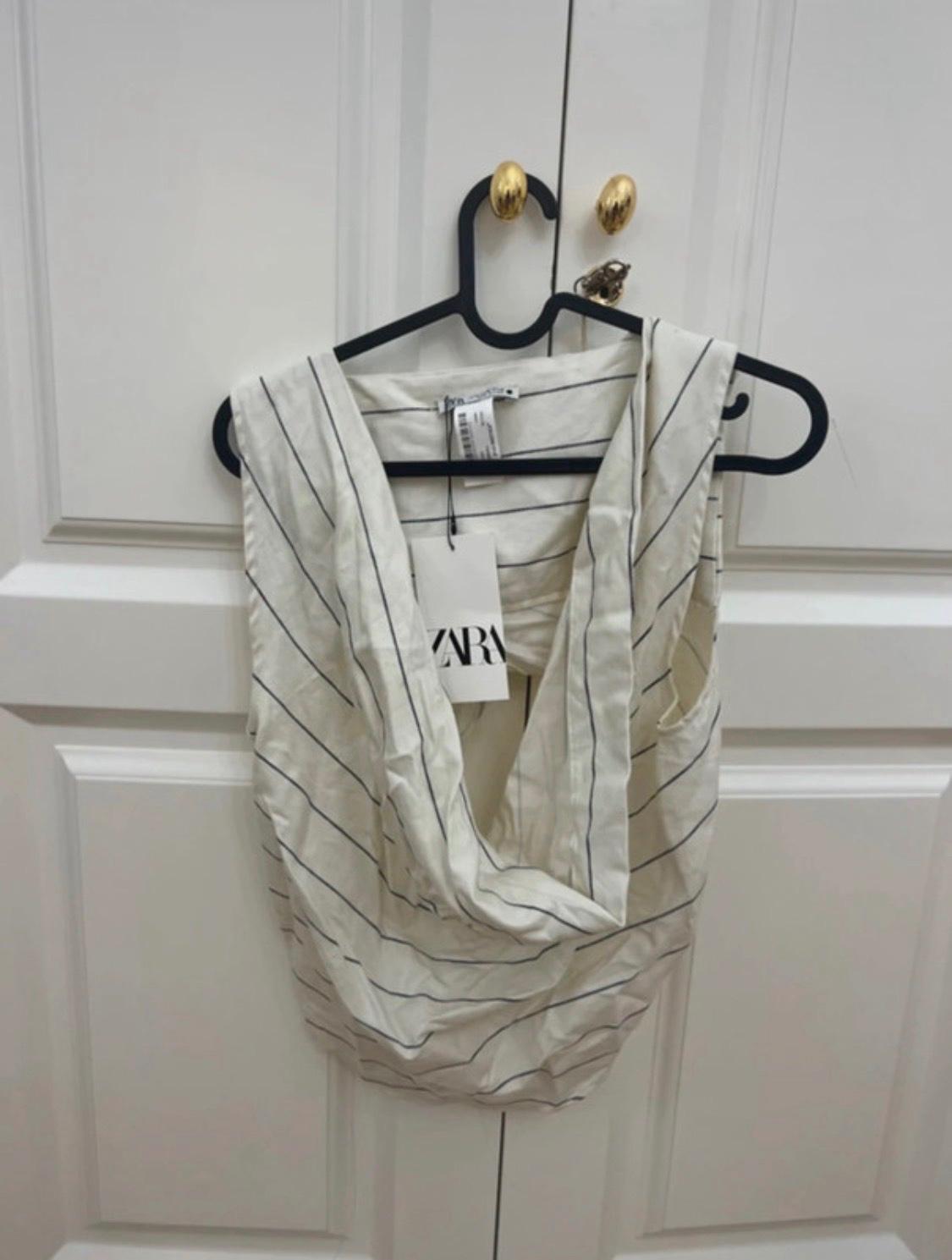 White top with dark stripes and a cowl neck hanging on a black hanger from the gold knob of a white door. Label with the text ZARA.