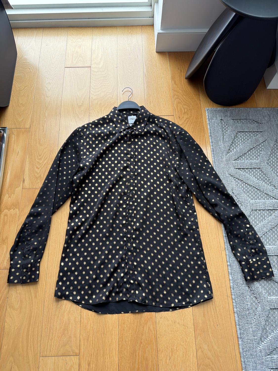 Black shirt with gold polka dot print on a light wood floor. Black hanger visible at the top.