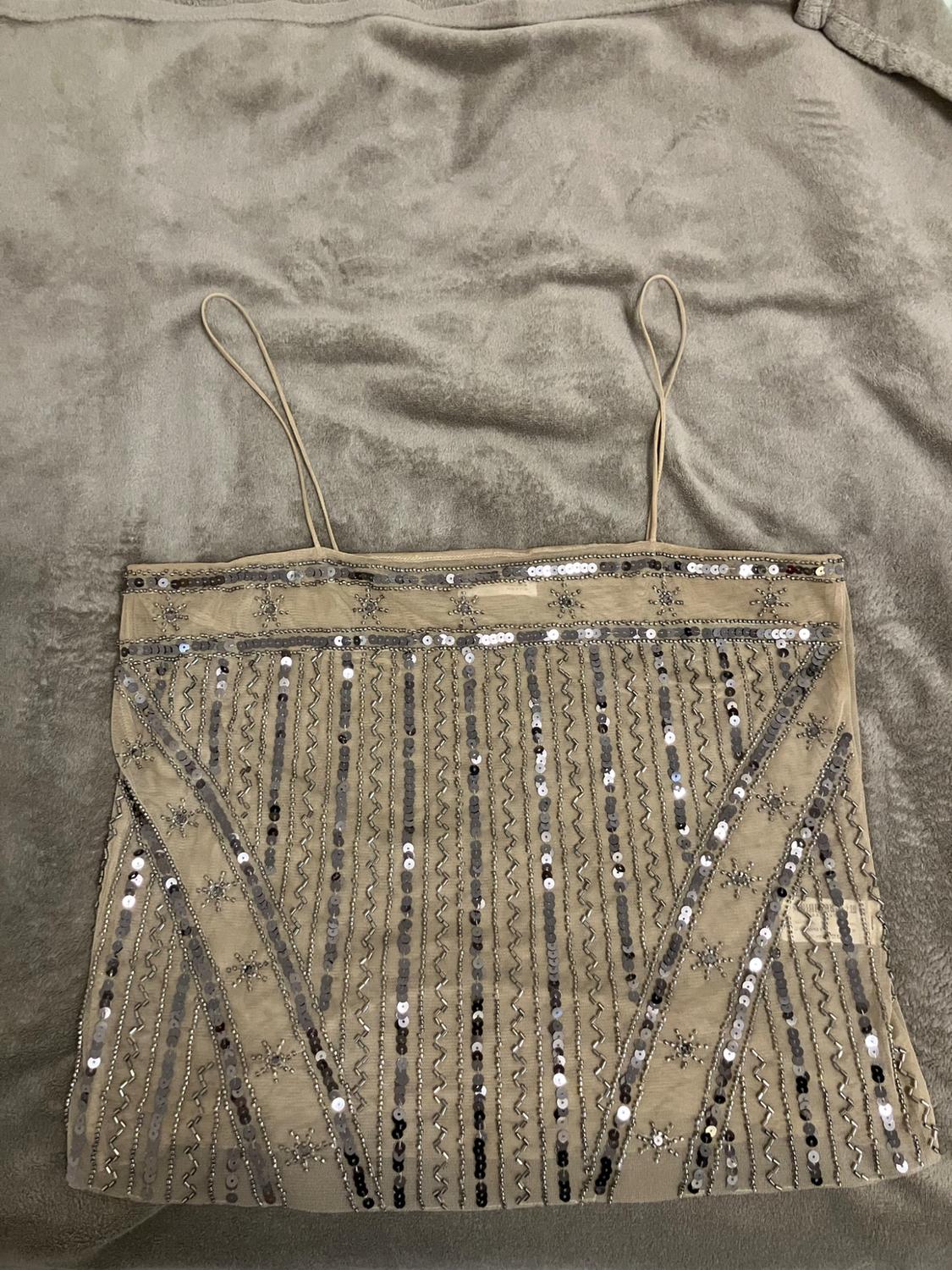 Top beige semitransparente de tirantes finos, decorado con lentejuelas plateadas en líneas y estrellas, sobre una manta suave.