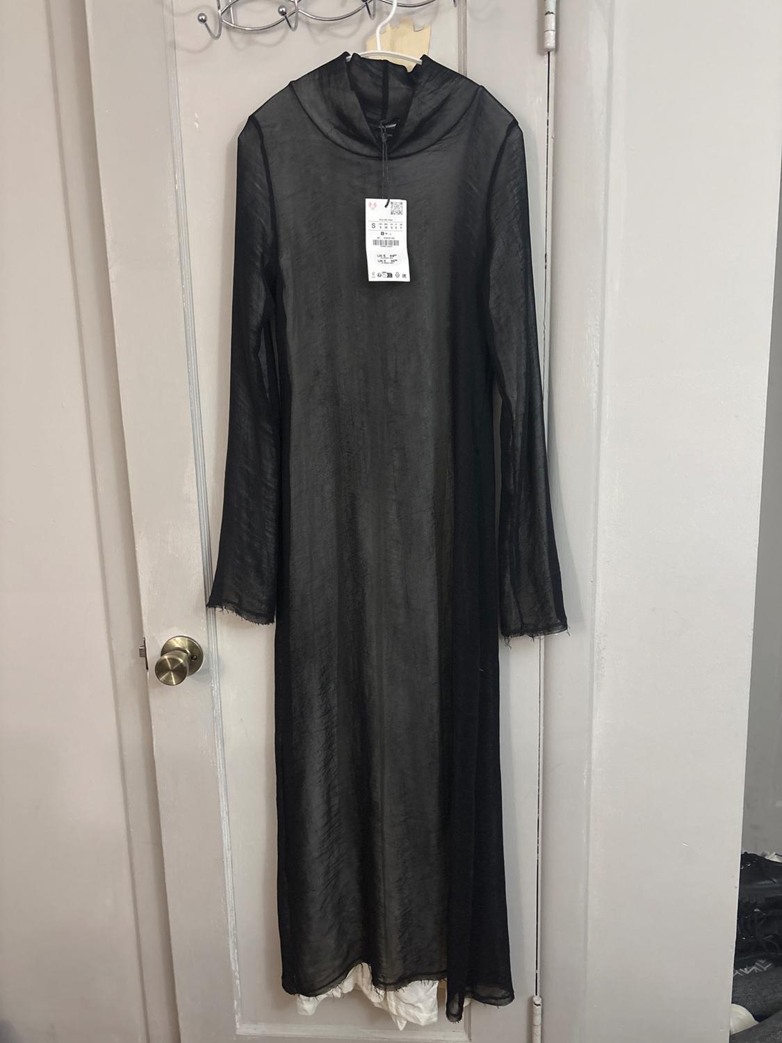 Long, semi-transparent black dress with long sleeves and a label, hanging on a white door.