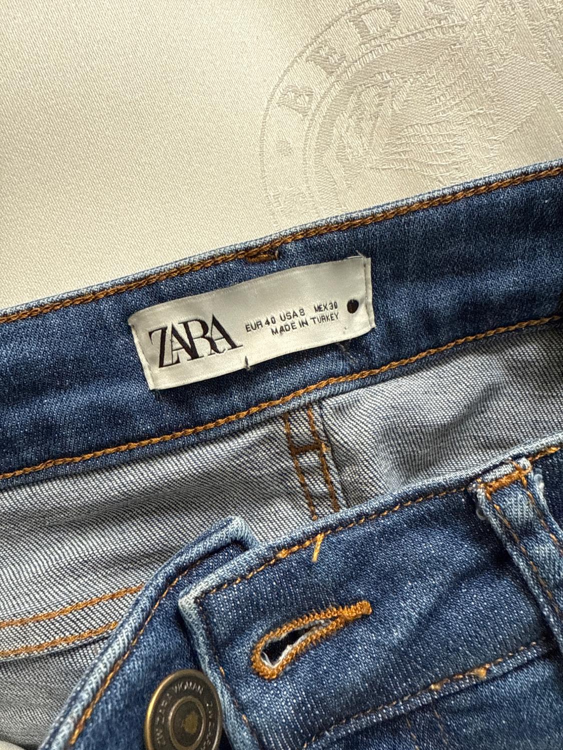 Blue jeans label with orange stitching. Visible text: "ZARA, EUR 40 USA 8 MEX 30, MADE IN TURKEY". Light background with embossing.