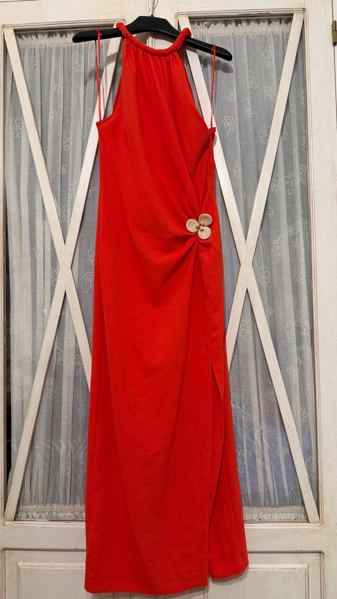 Red maxi dress with a halter neck, side ruching with a flower detail and a slit, hanging in front of a door with glass panels and white curtains.