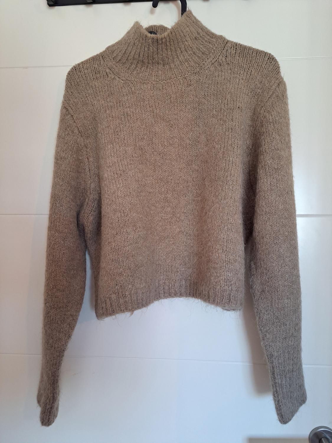 Beige knitted roll-neck jumper, hanging on a hanger against a white background. Simple design and soft texture.