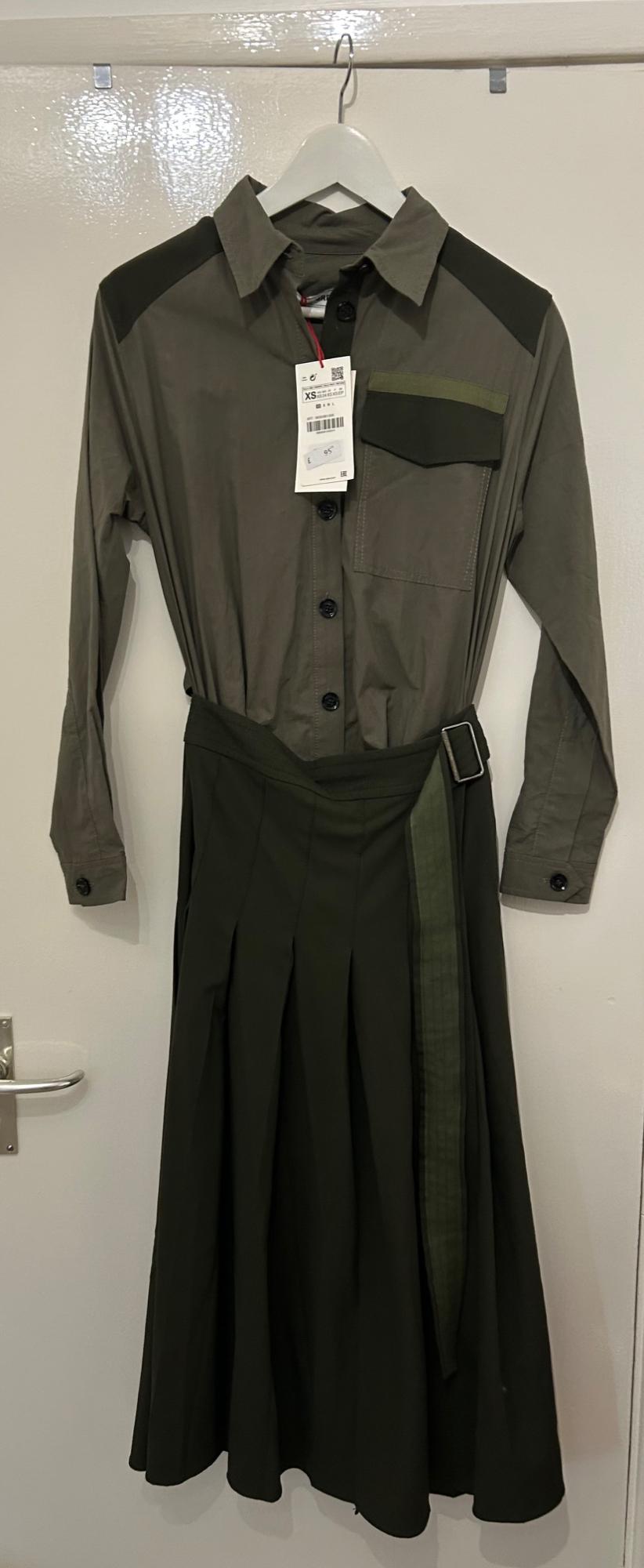 Olive green dress with black buttons and belt, hanging on a white hanger. Visible tag with size XS and price.