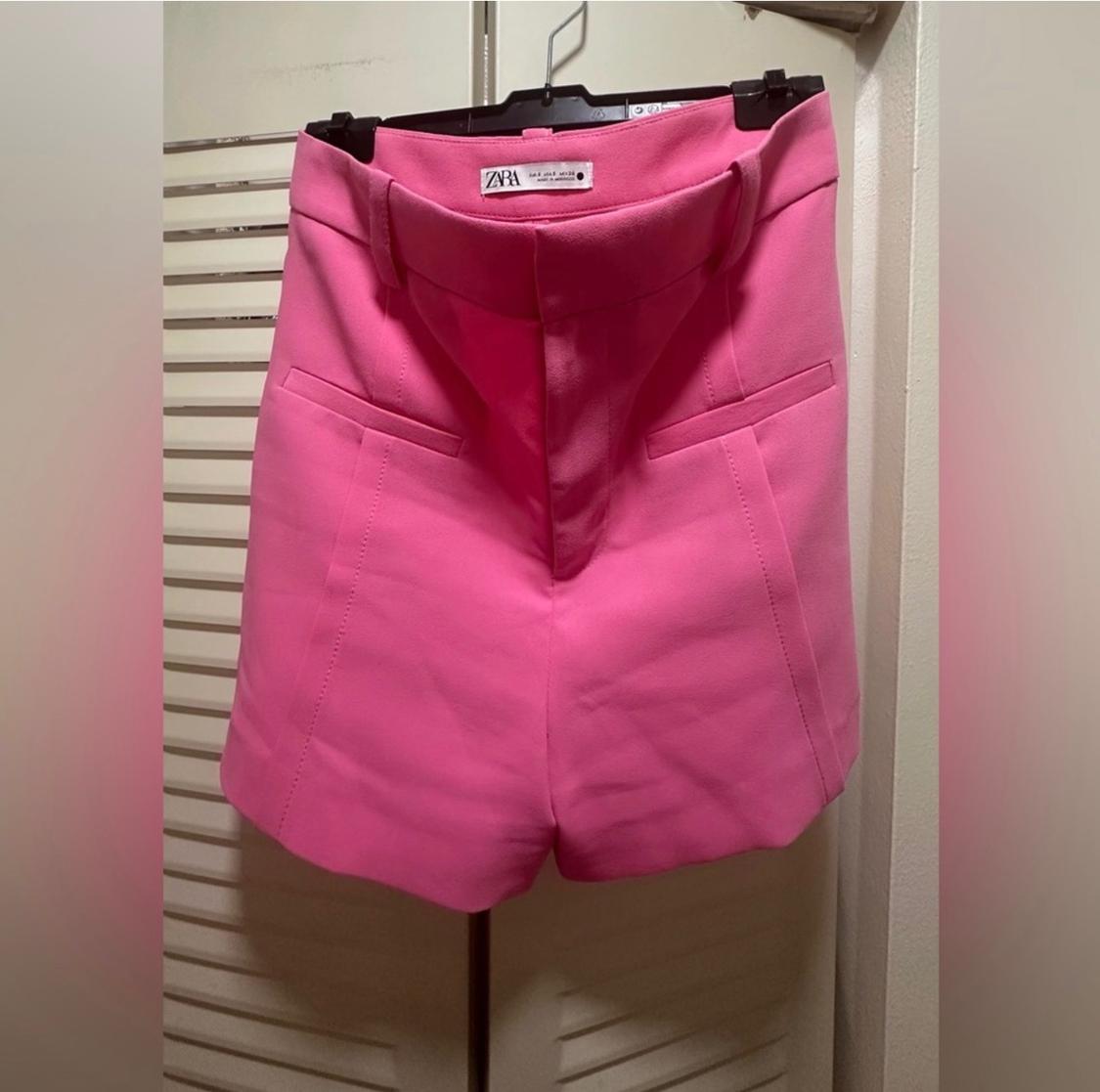 Bright pink shorts hanging on a hanger, with front pockets and a visible label on the waistband that reads "ZARA". White door background.