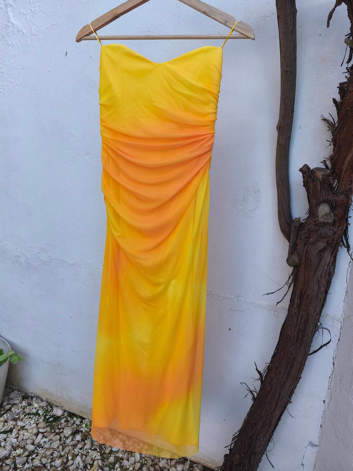 Long yellow and orange dress with spaghetti straps, hanging on a wooden hanger against a white wall, next to a dry branch.