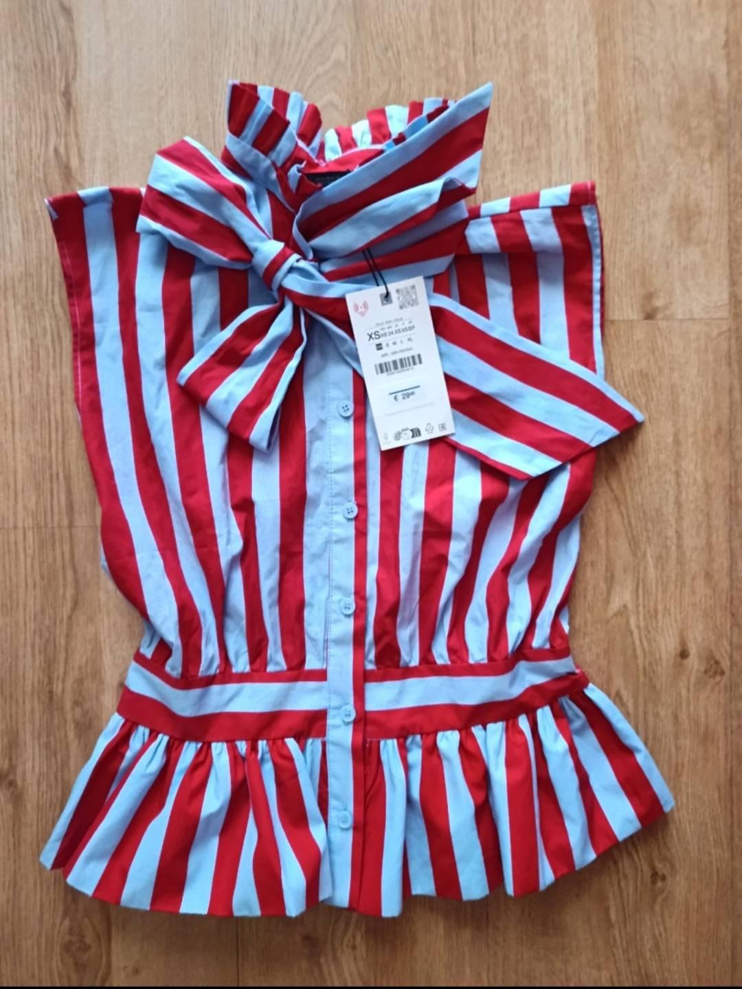 Overhead view of a button-down blouse with red and light blue stripes, neck tie and flared hem, on wood. Label with text XS and € 29.