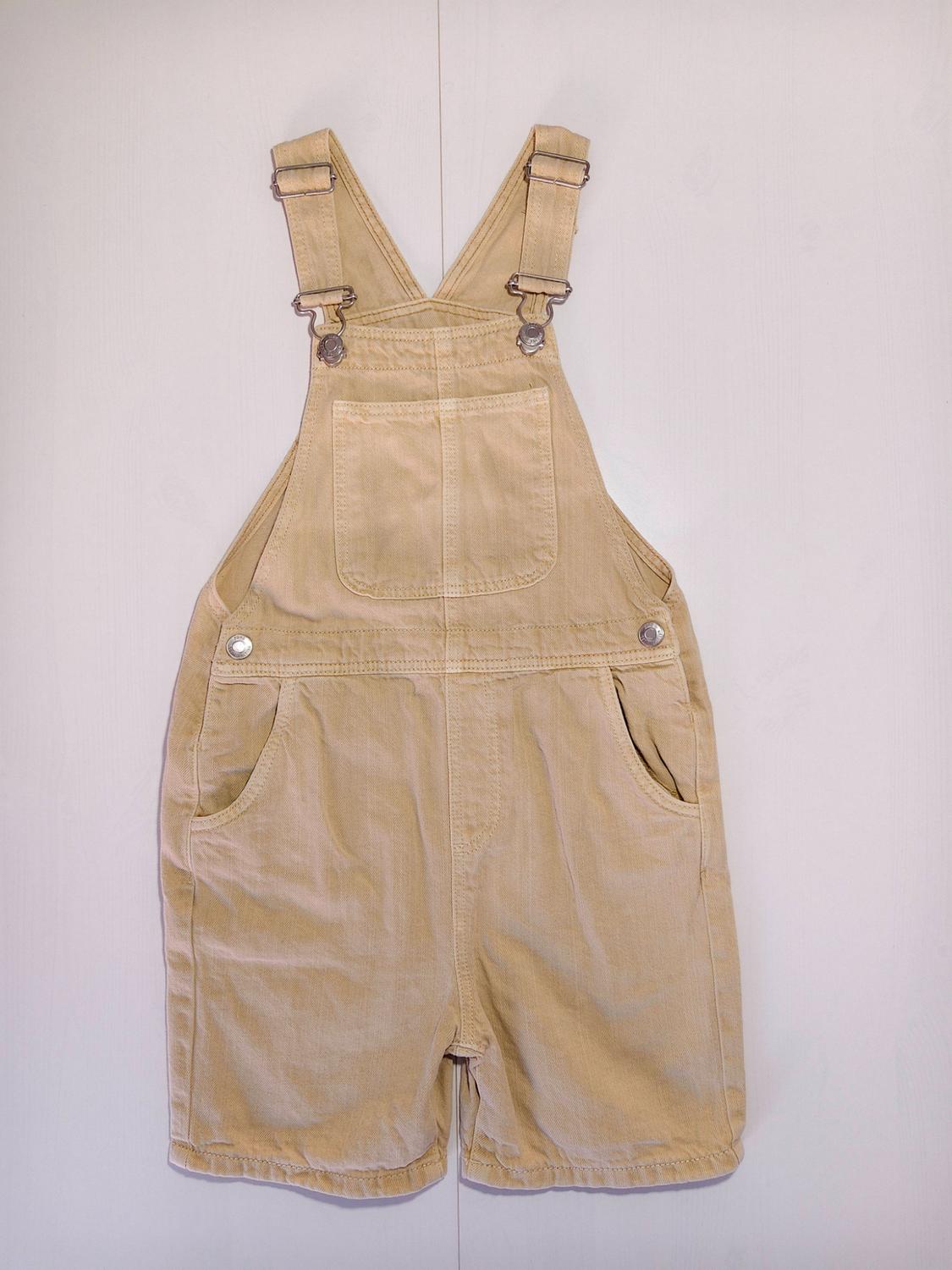 Beige short dungarees with a front pocket, side pockets, and straps with metal fastenings, set against a light background.
