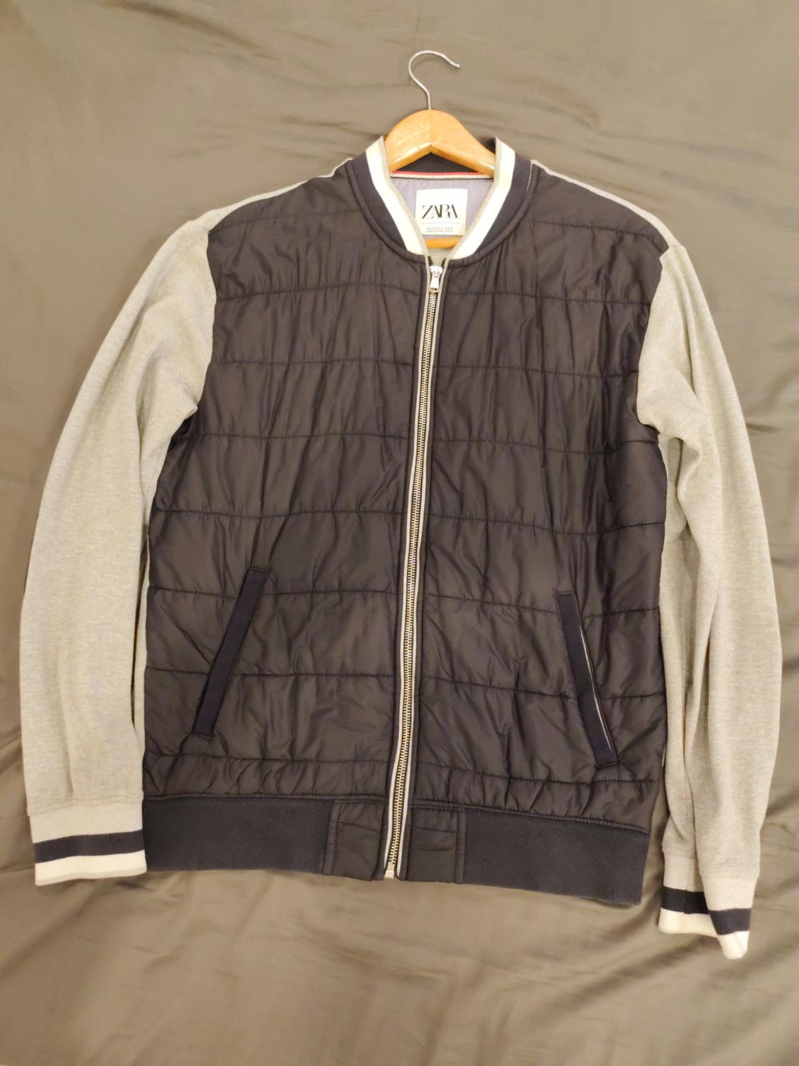 Μπουφάν τύπου bomber με ετικέτα ZARA σε κρεμάστρα. Διαθέτει σκούρο καπιτονέ σώμα, γκρι μανίκια και κλείσιμο με φερμουάρ στο μπροστινό μέρος.