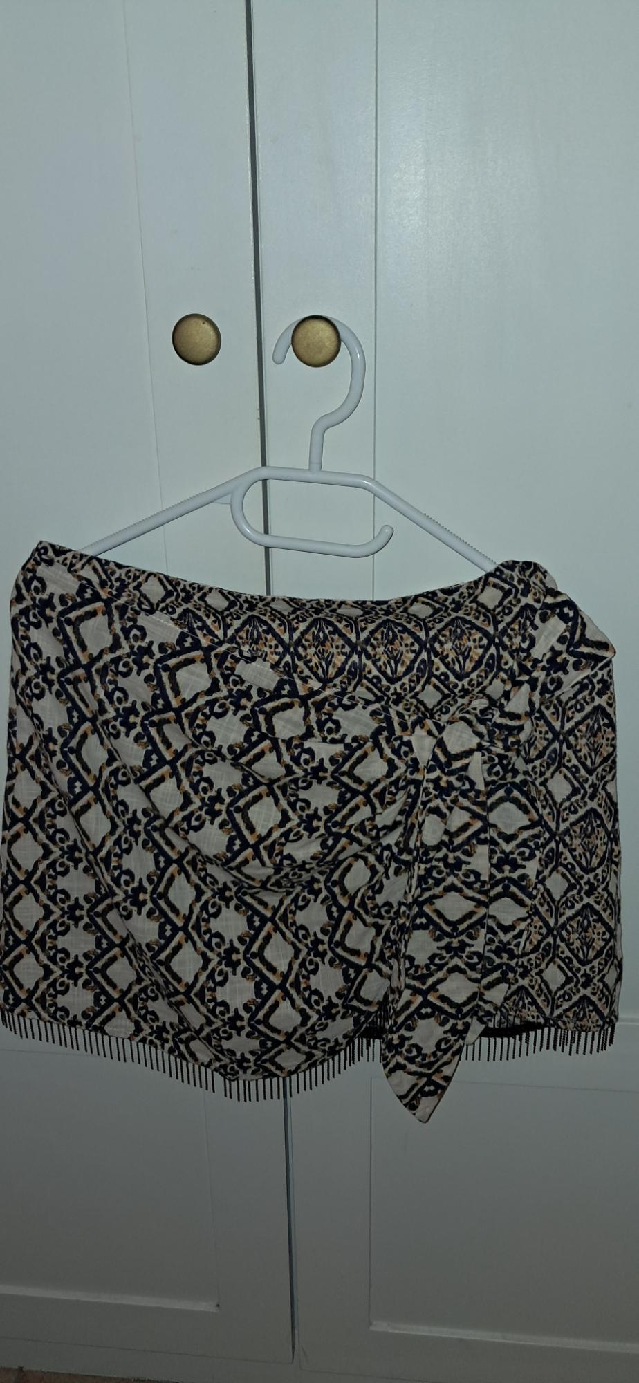 Short skirt with a geometric print, side knot detail and fringing at the hem, hanging on a hanger over a white wardrobe.