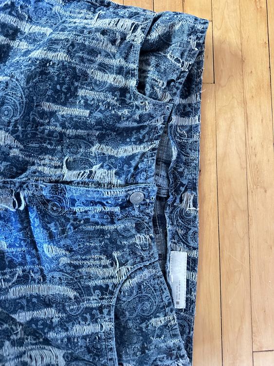 Blue denim jeans with a distressed design and an abstract pattern on a light wood floor. Visible tag on the inside.