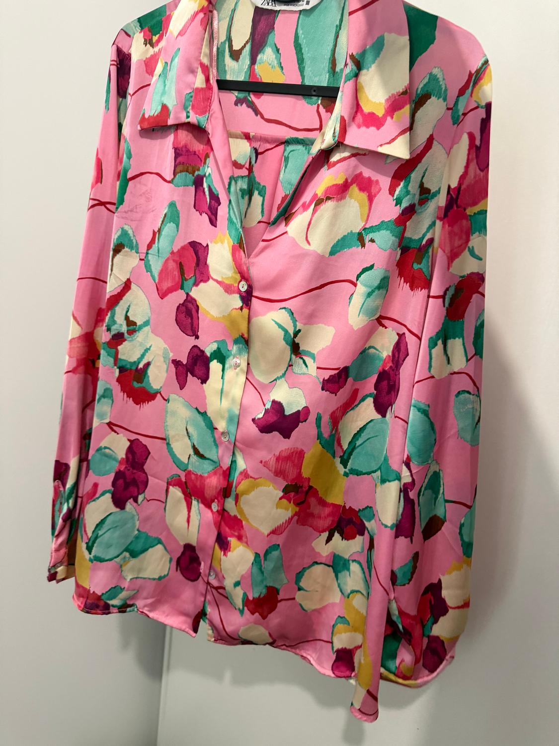 Women's shirt with a floral print in pink, green, and yellow tones, hanging on a hanger against a white background.