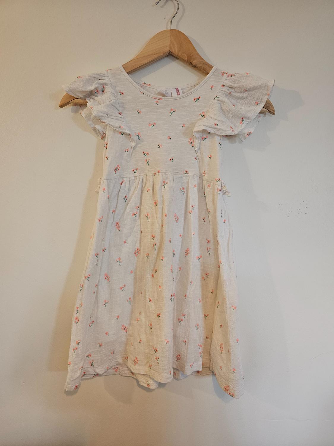 White children's dress with a small pink floral print and ruffled sleeves, hanging on a wooden hanger.