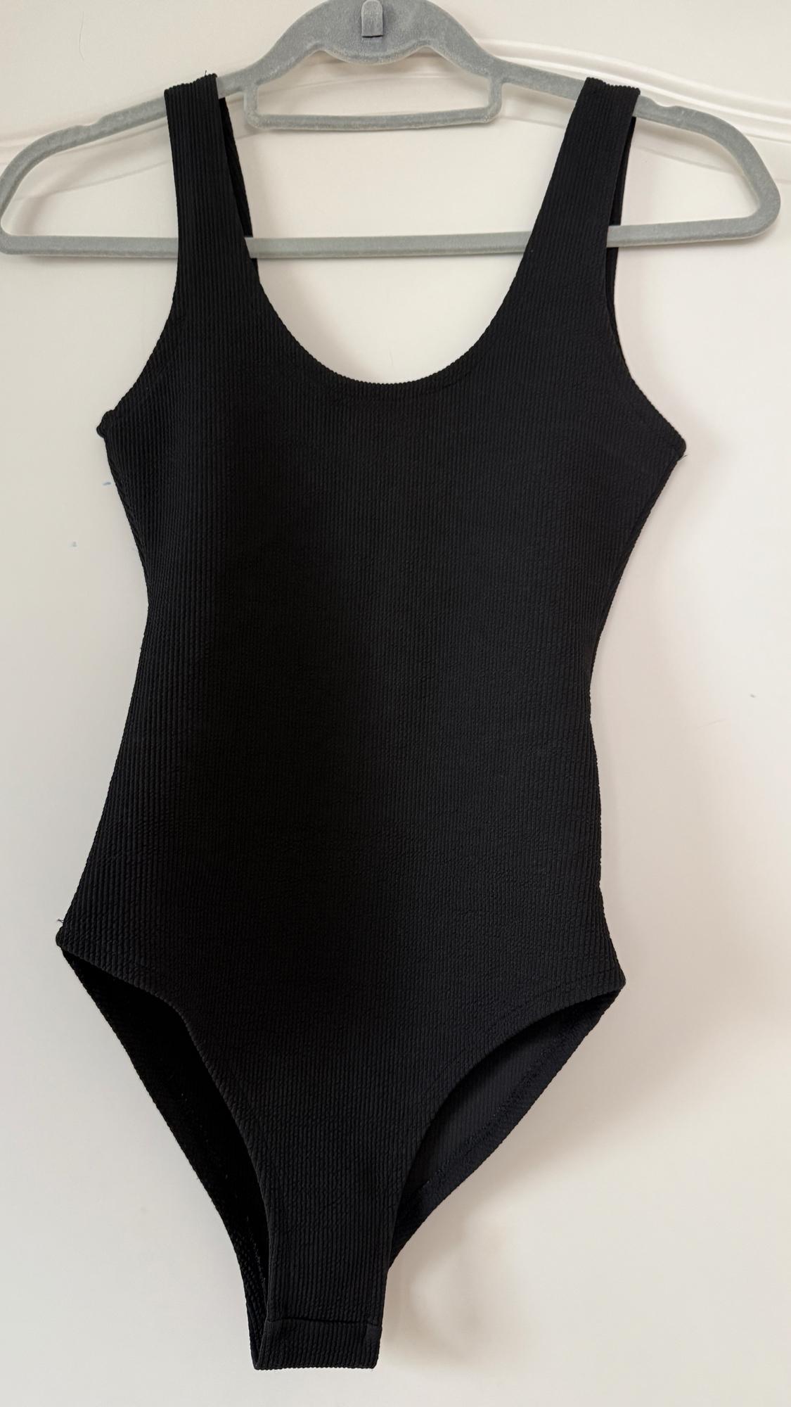 Black thick-strap bodysuit hung on a grey hanger on a white background. Ribbed fabric.