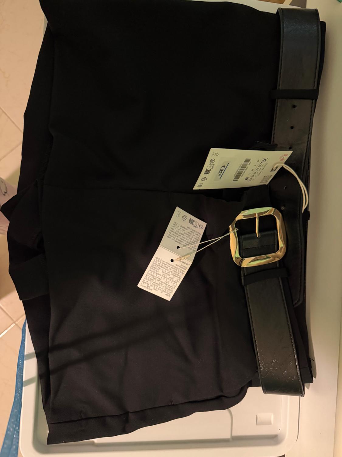Black trousers, folded, with a black belt and gold buckle on a white surface. Tags hanging from the belt.