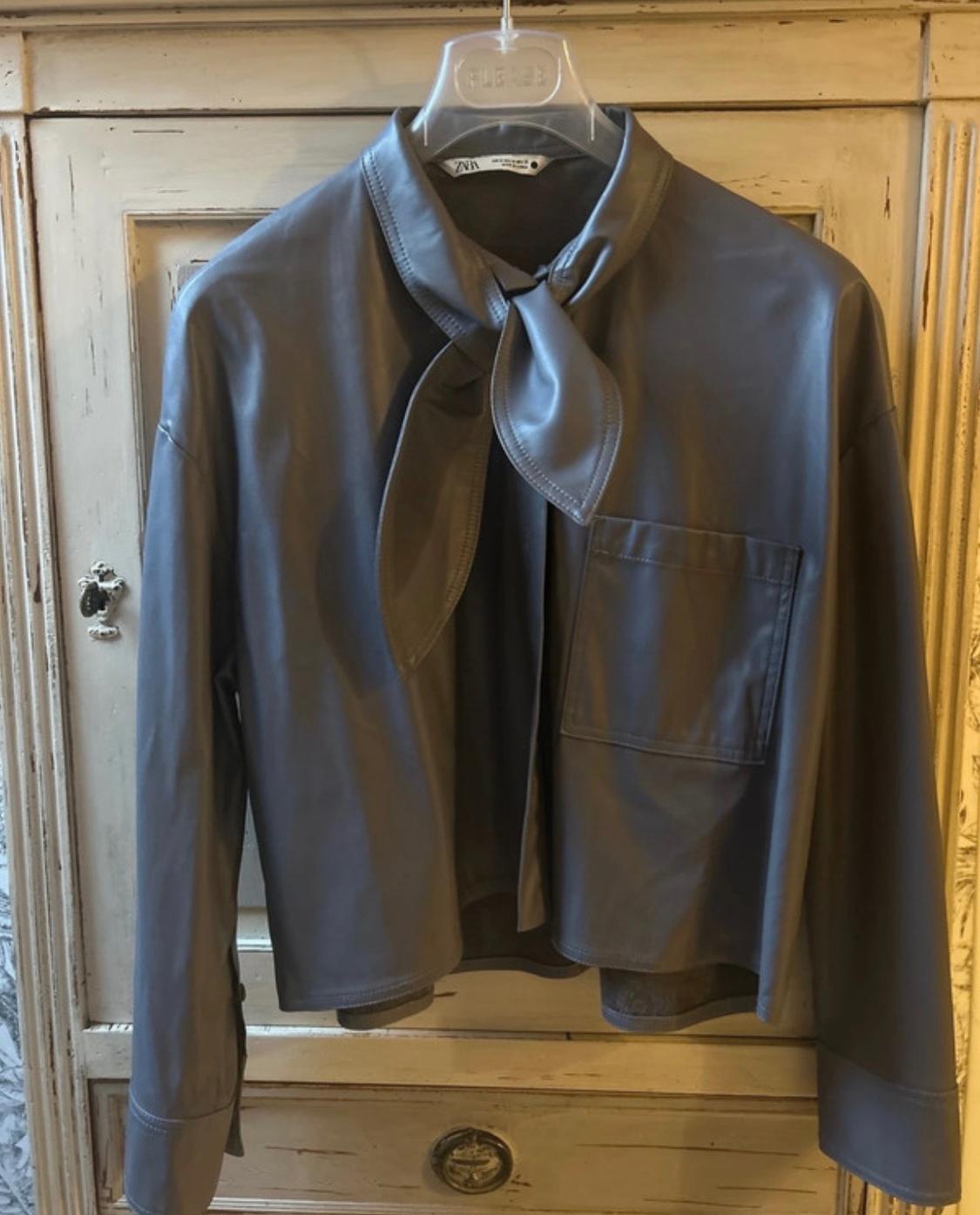 Grey jacket hanging on a clear hanger, with a bow at the neck and a front pocket, on a light wood cabinet.