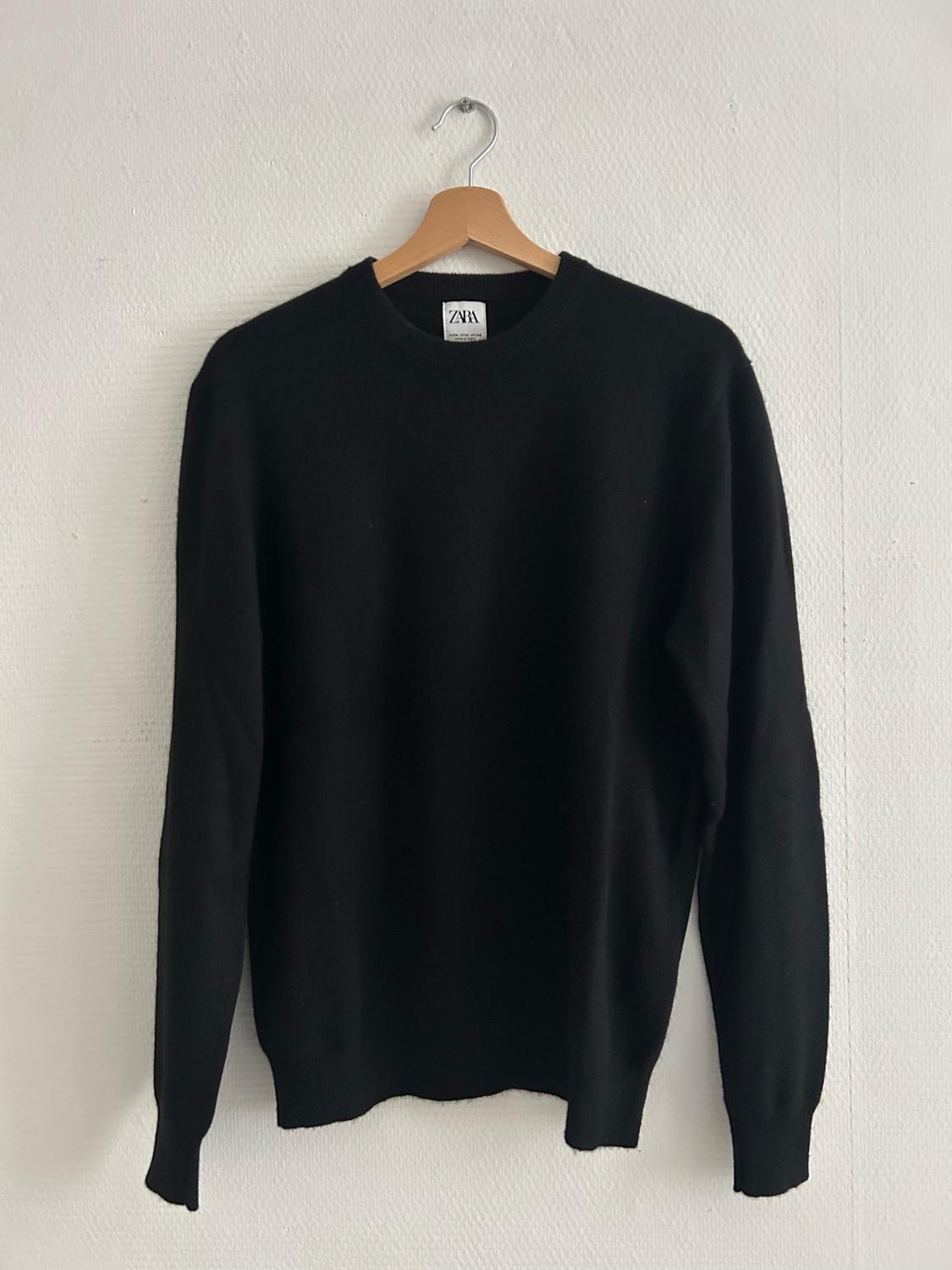 Black long-sleeved jumper hanging on a wooden hanger. Visible label with the text "ZARA". White background.