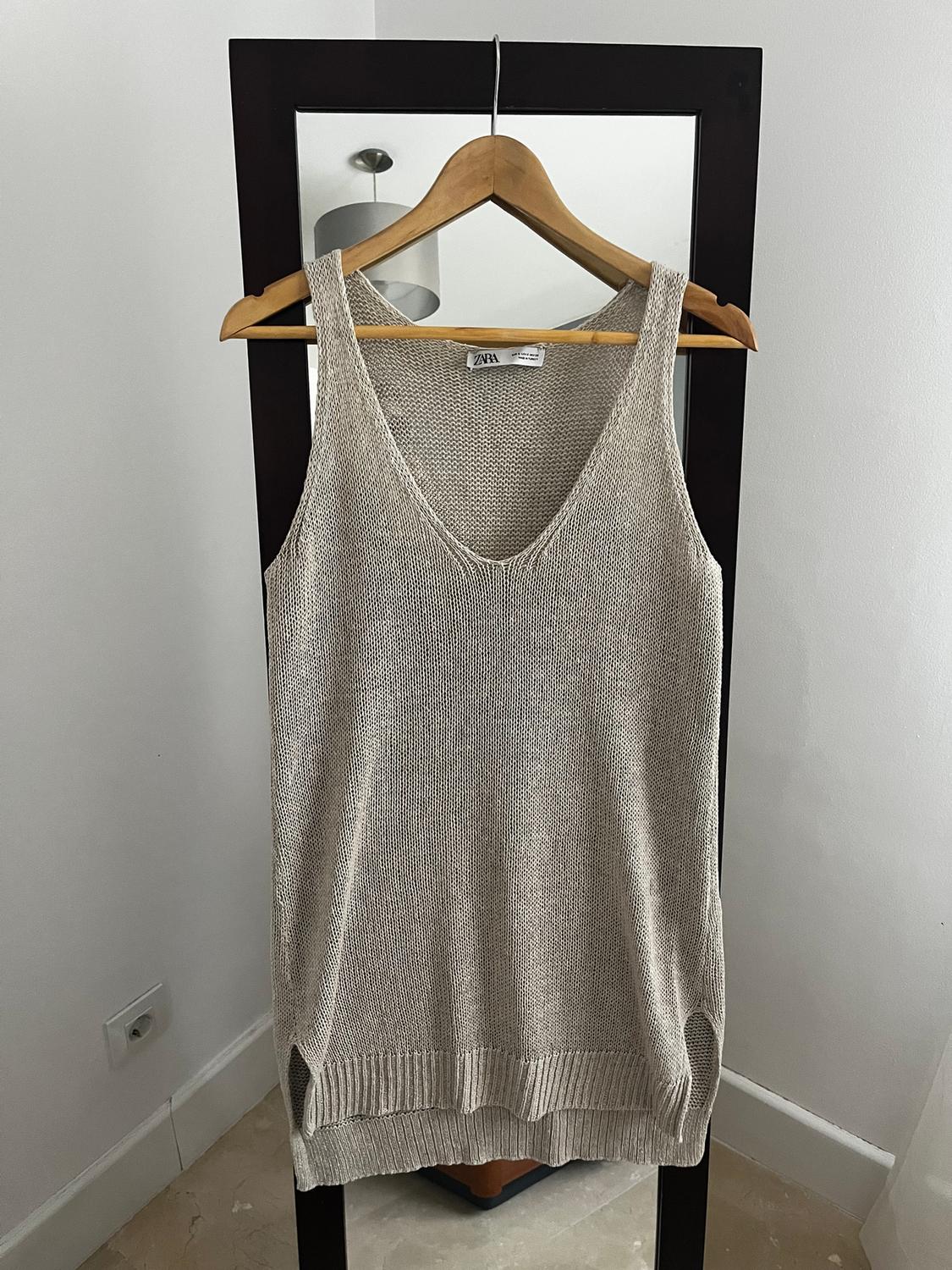 Sleeveless beige jumper hanging on a wooden hanger in front of a mirror. Visible label with the brand "Zara". Minimalist white background.