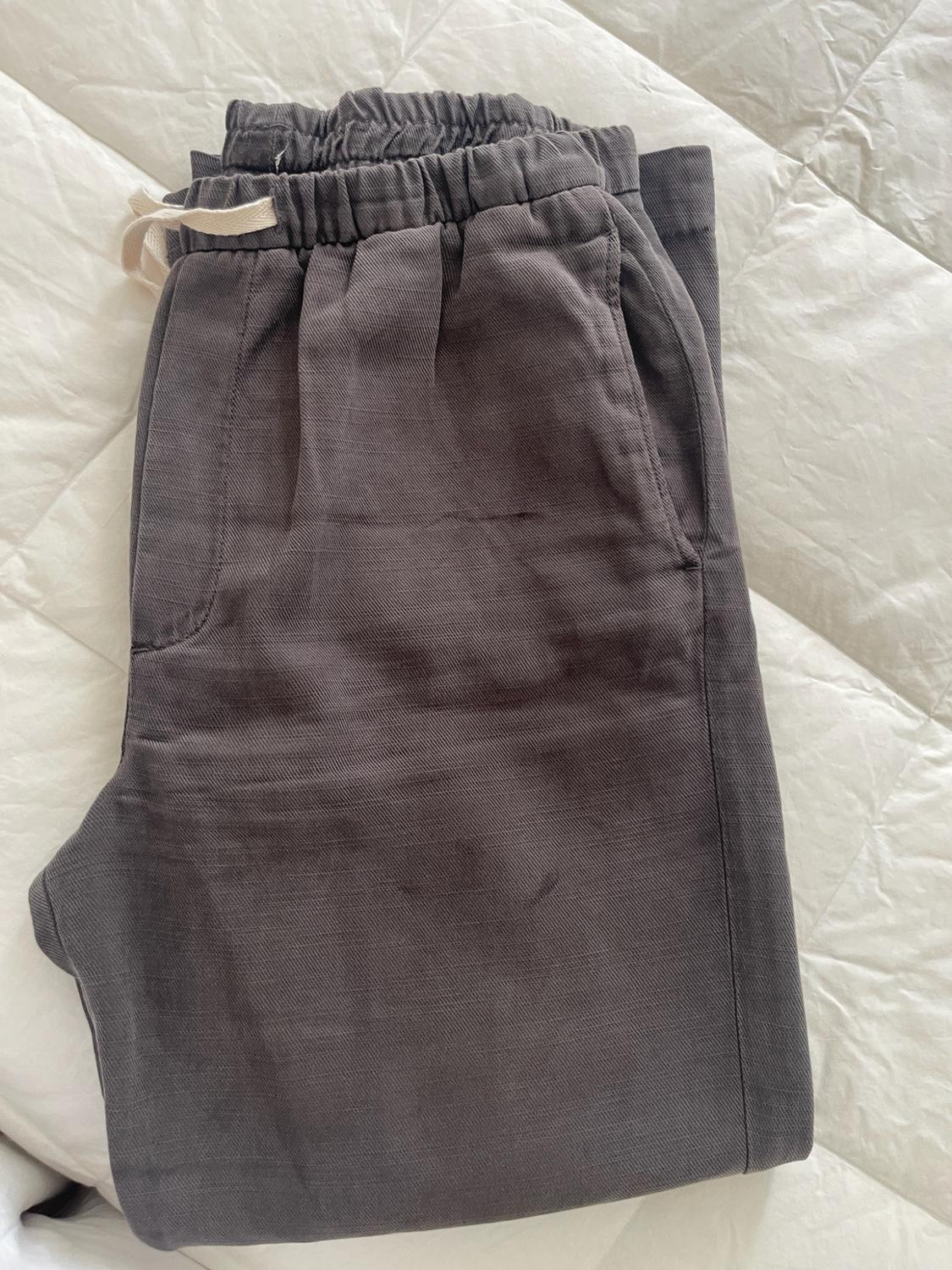 Dark grey trousers folded on a white bed. Casual design with an elasticated waist, light-coloured drawstring and side pockets.