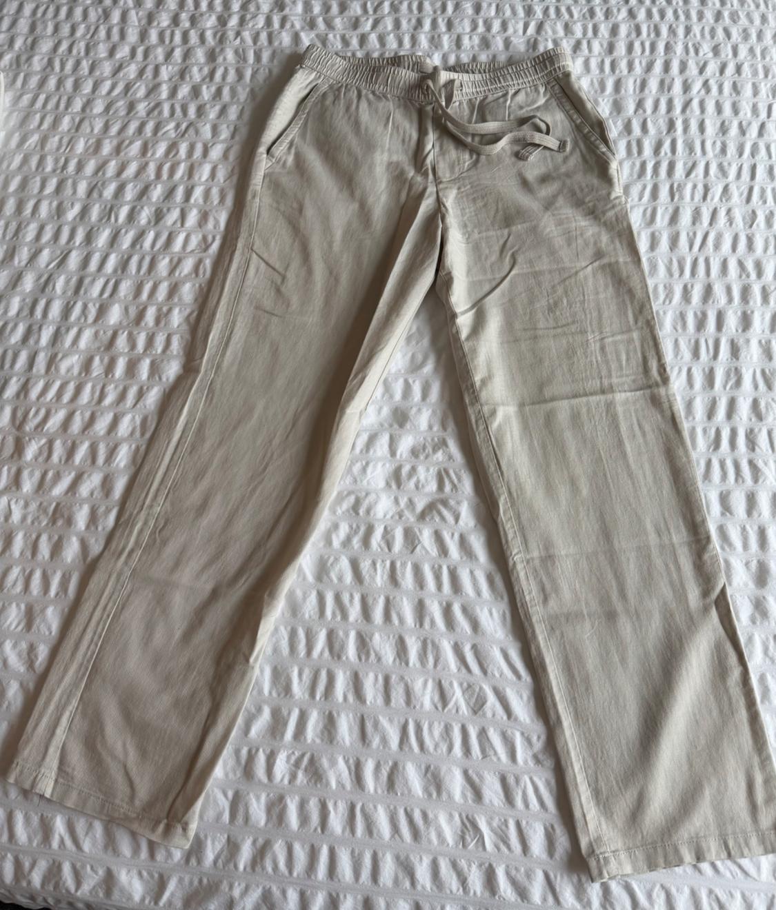 Light beige casual pants with an elastic waistband and adjustable drawstring, displayed on a textured white bedspread.
