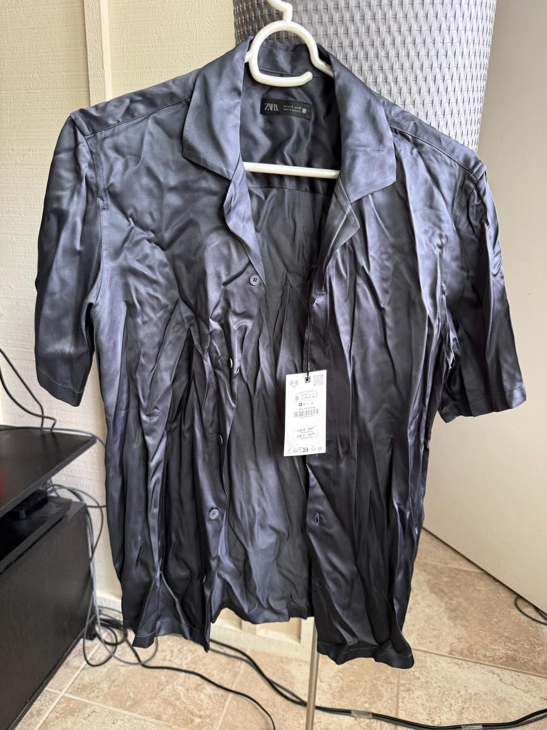 Crumpled dark short-sleeved shirt on a white hanger. Label with ZARA text on the collar and price tag. Indoor setting.