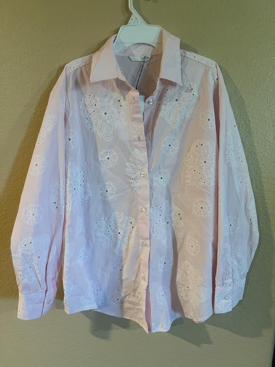 Light pink shirt with white floral embroidery, hanging on a white hanger against a beige wall.