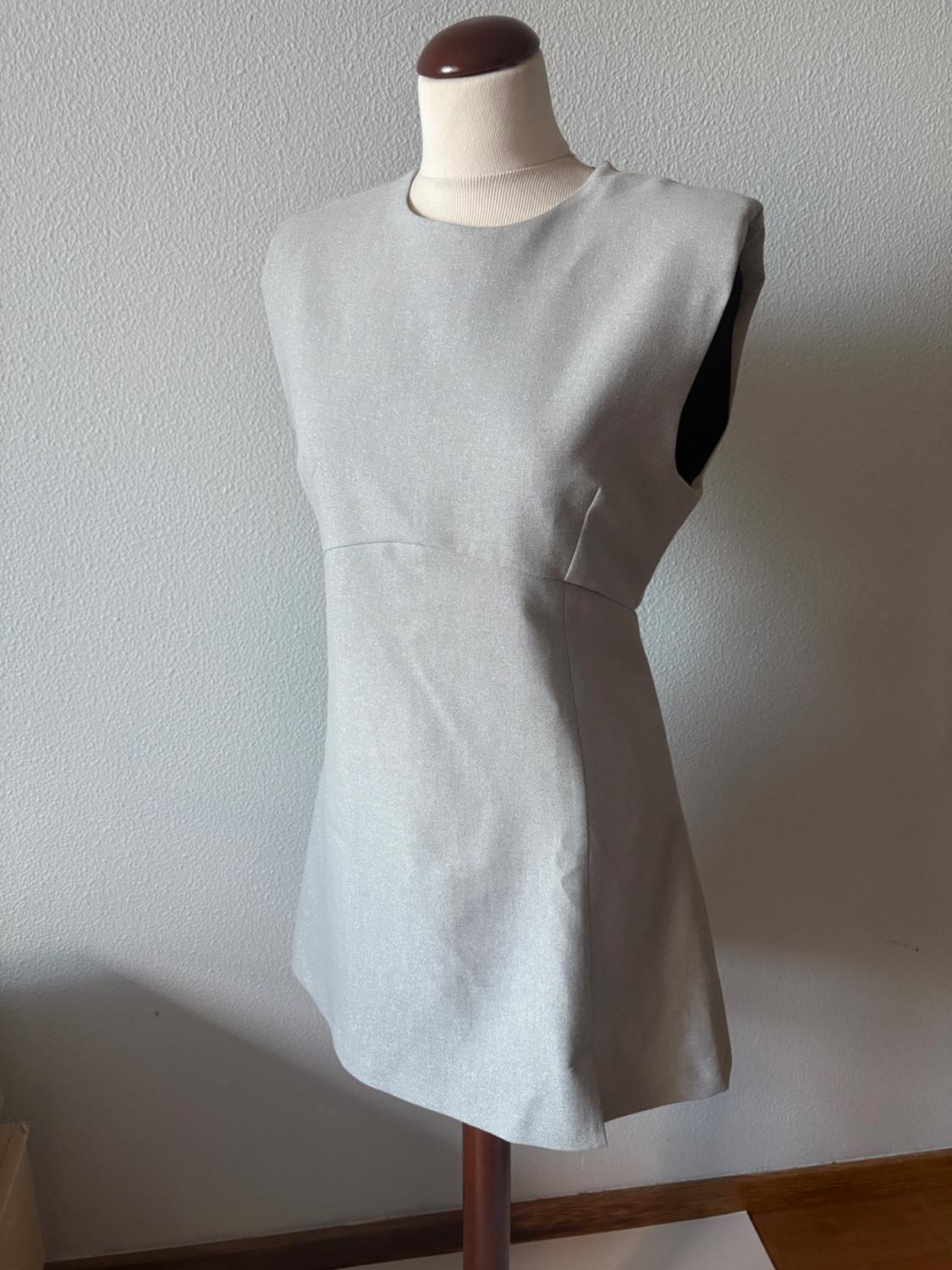 Mannequin wearing a light grey sleeveless mini dress with a subtle shimmer. Round neck design in front of a textured white wall.