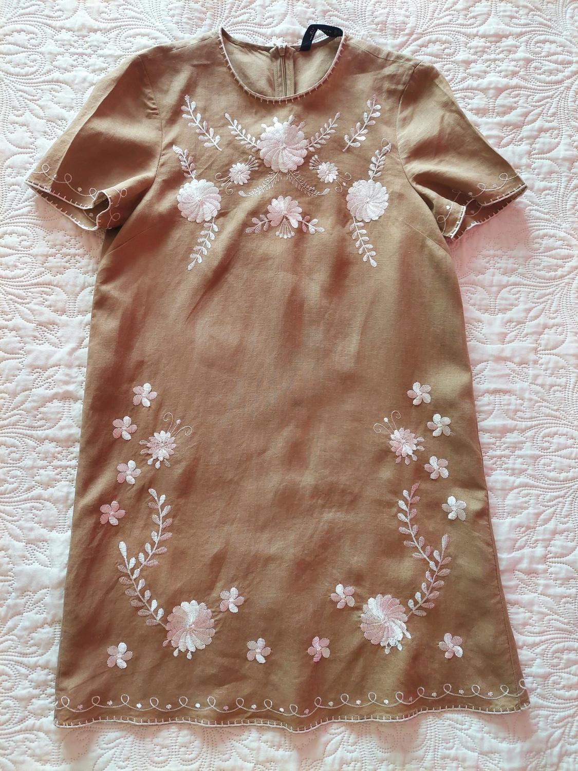 Short, light brown, short-sleeved dress with white floral embroidery, laid out on a white textured bedspread.