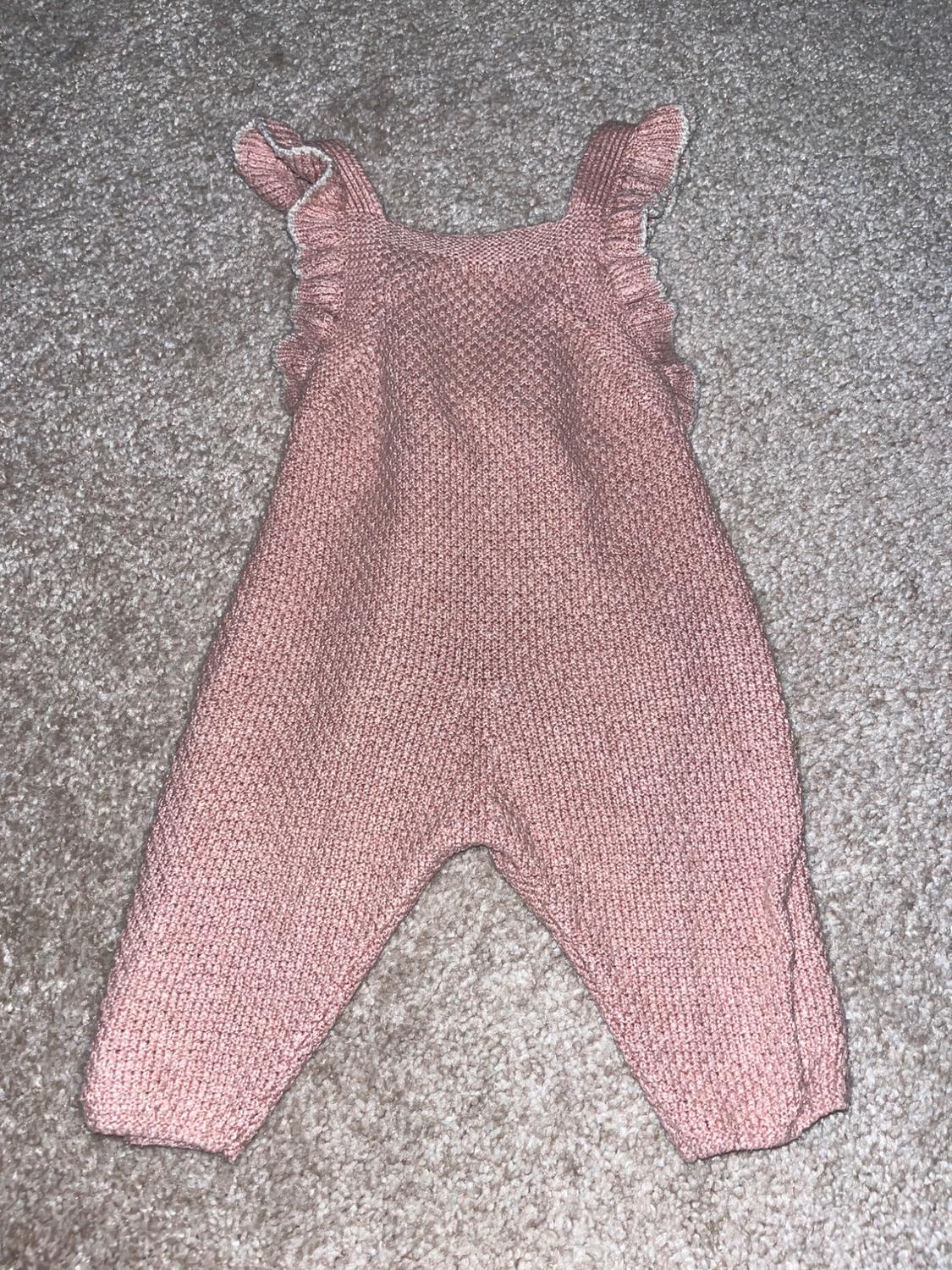 Pink knitted baby romper with straps and ruffles, on a grey rug.