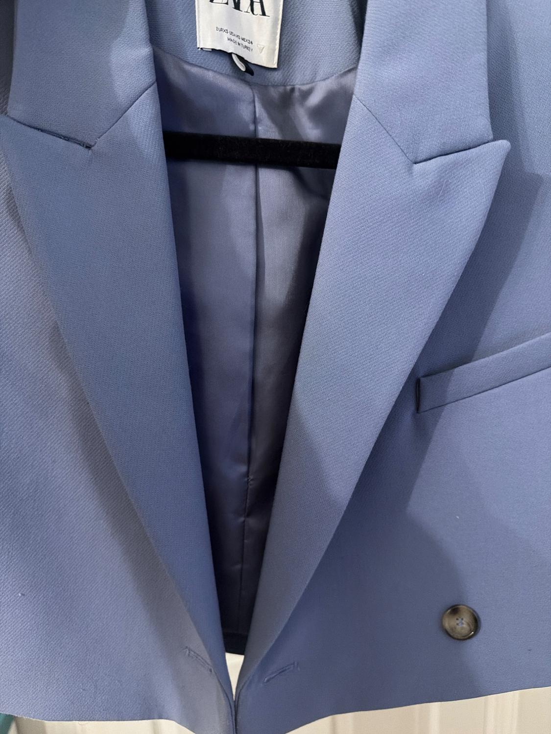 Light blue blazer hanging on a hanger, with wide lapels and a visible button. Internal tag with partially visible text.