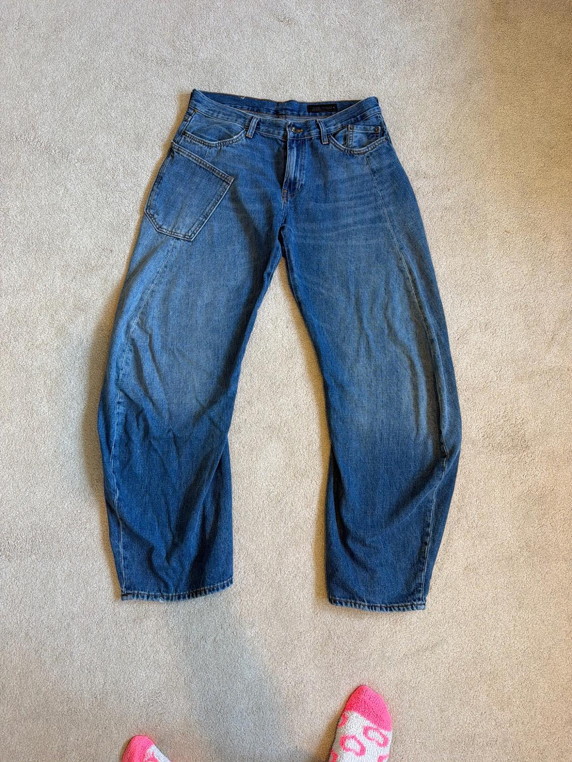 Wide blue jeans with a patch pocket on the thigh, over a light-coloured carpet. Below, a few feet can be seen, with pink socks.