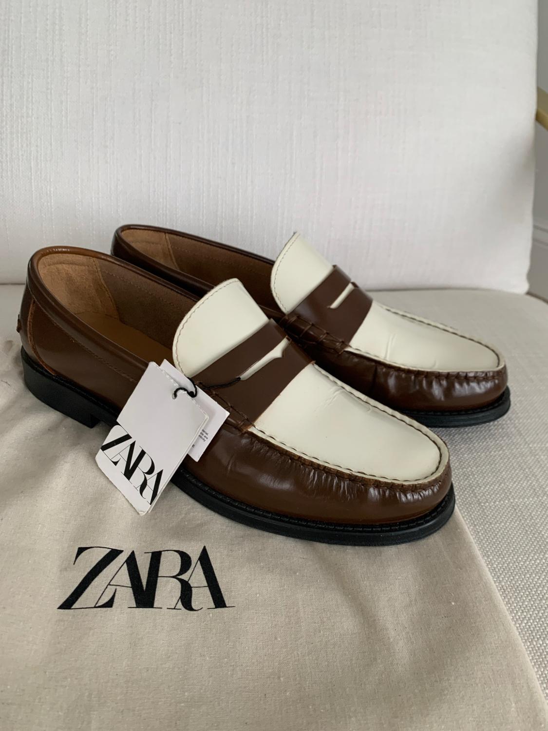 Brown and white loafers with a ZARA label, resting on a fabric bag bearing the text ZARA against a light background.