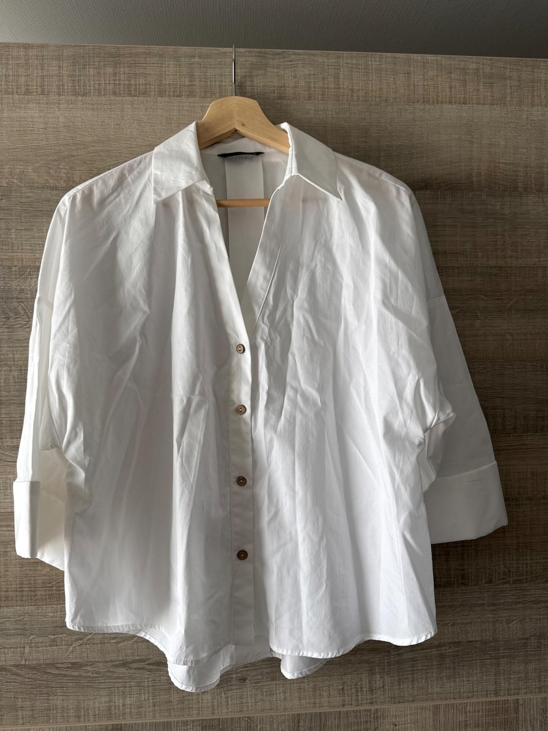 White long-sleeved shirt with brown buttons, hanging on a wooden hanger against a light wood background.