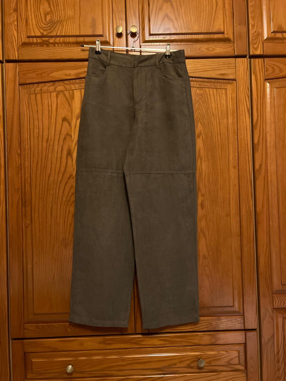 Dark long trousers with a horizontal seam on the leg, hanging on a clothes hanger in front of a wooden wardrobe.