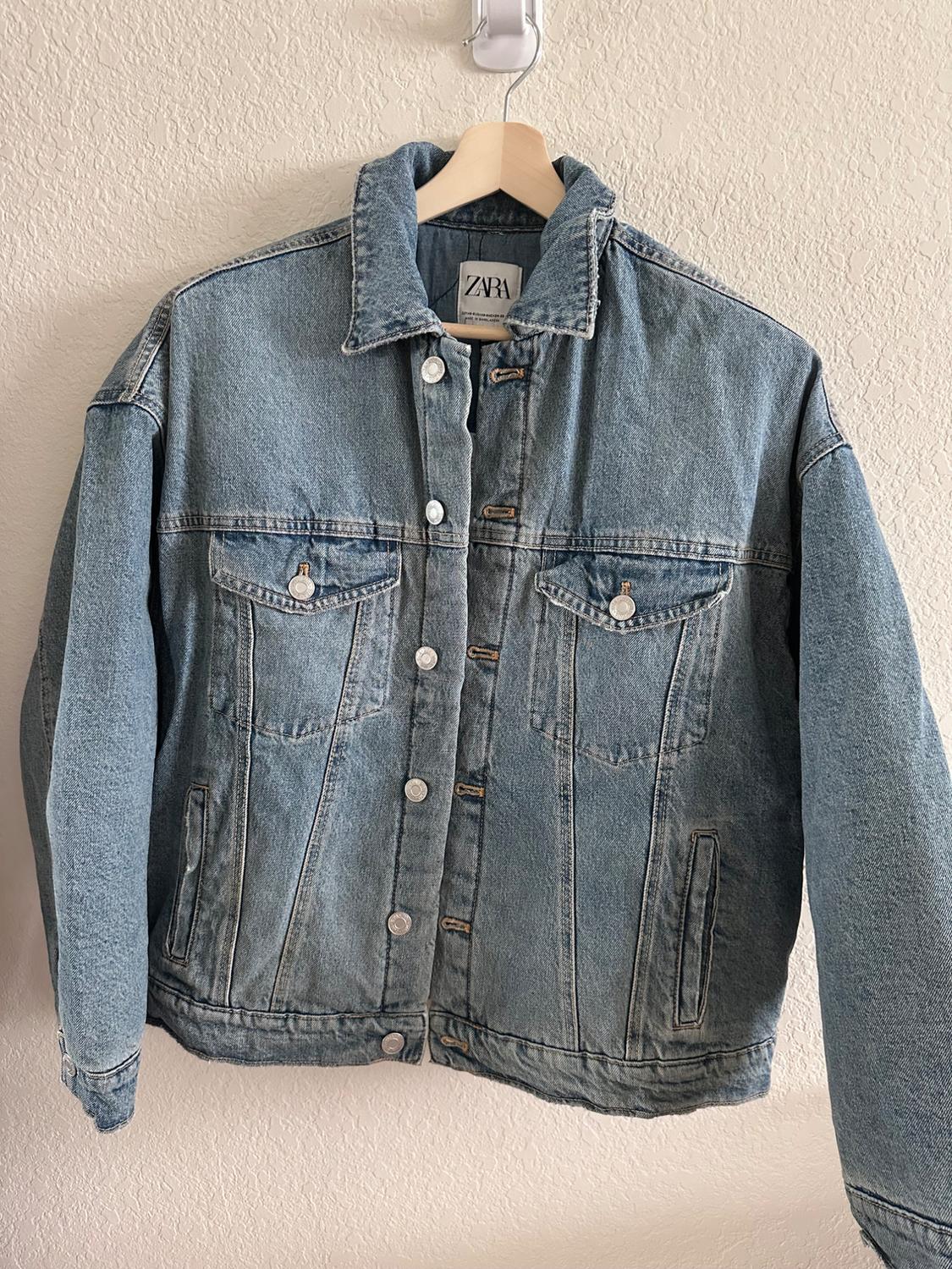Blue denim jacket hanging on a wooden coat hanger, with metal buttons and front pockets. Visible label with the text "ZARA".