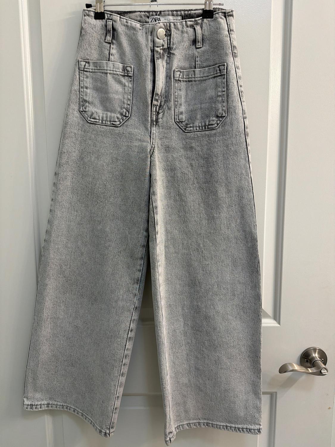 Light gray wide-leg denim pants hanging on a white door. Large front pockets and visible Zara tag.