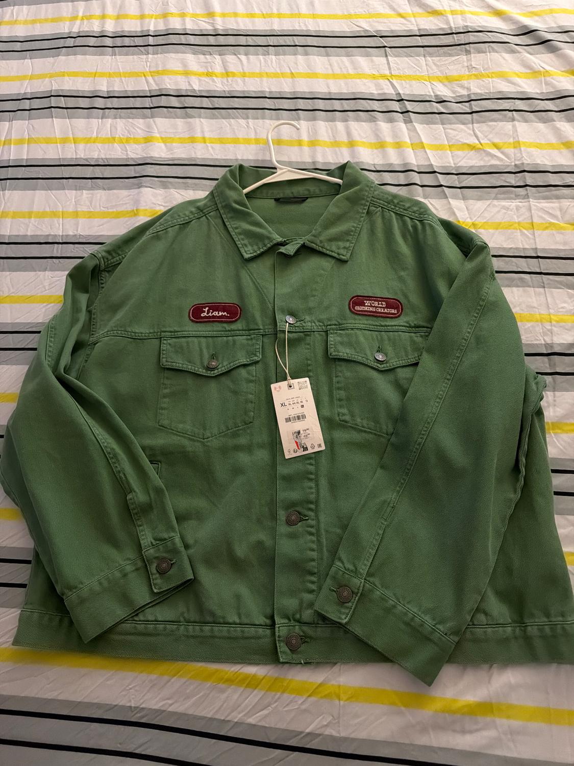 Green jacket with "Liam" and "World Changers" tags on a bed with grey and yellow striped sheets.