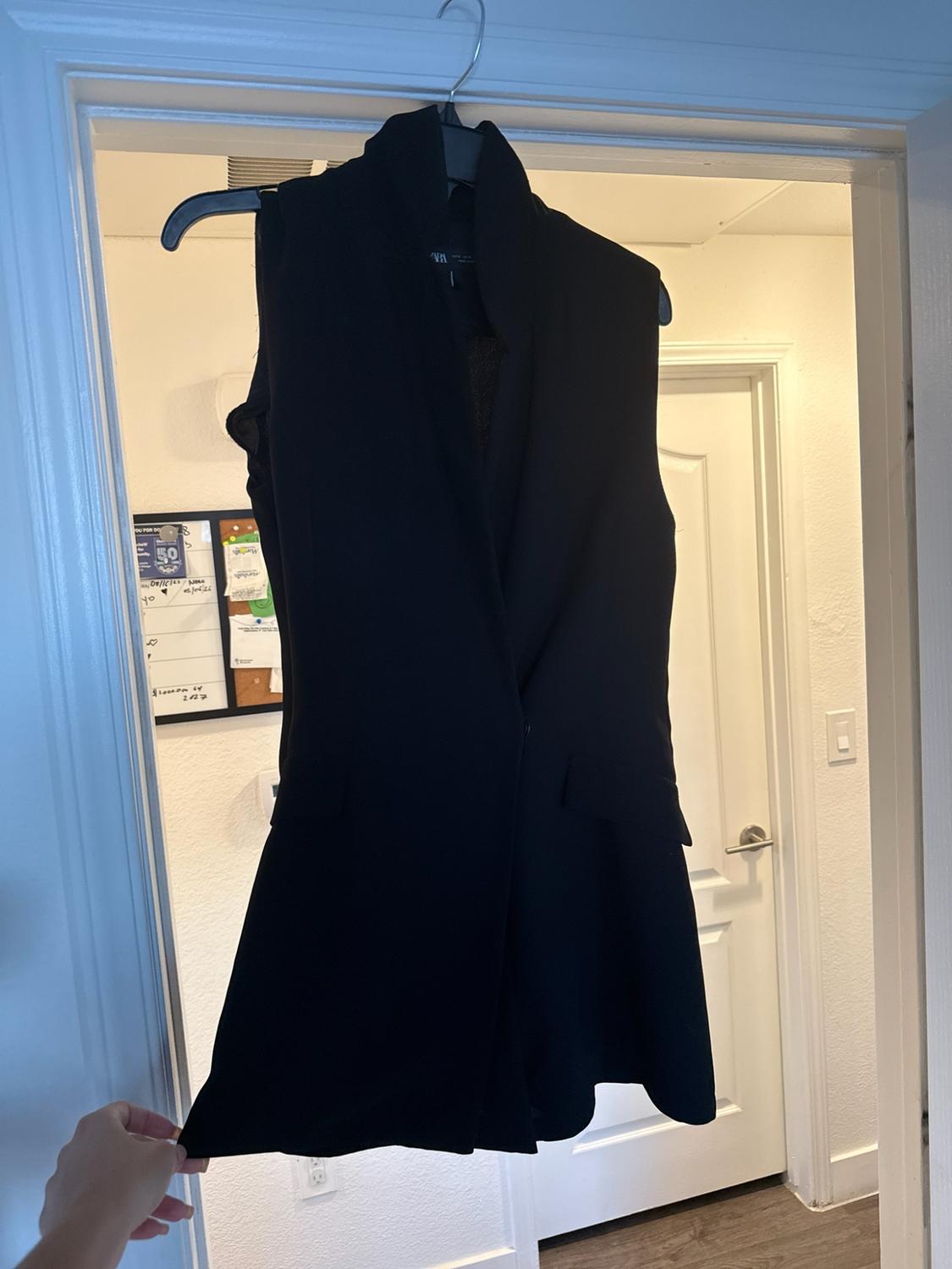 A black sleeveless vest hangs from a door. A hand holds the bottom of the garment. In the background, a room with white walls is visible.