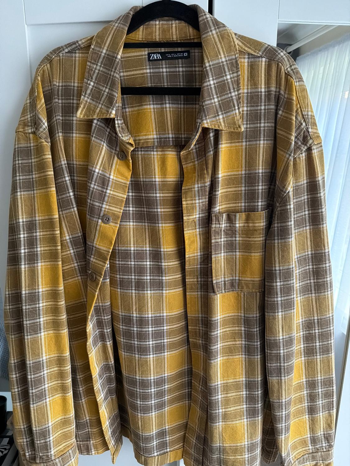 Brown and yellow checked shirt hanging on a hanger. Visible brand tag. White wardrobe and curtain in the background.