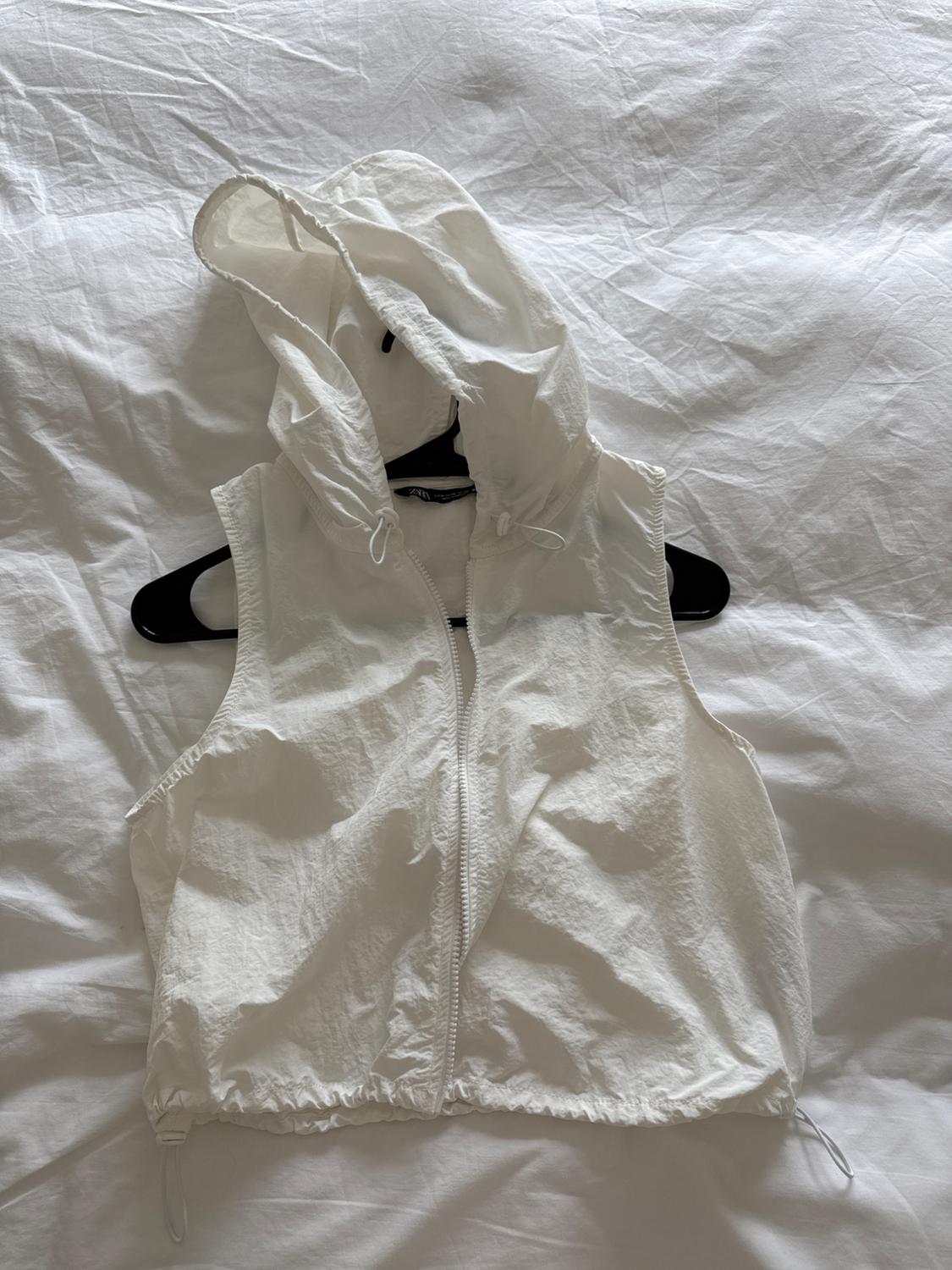 White hooded zip-up vest, hanging on a black hanger over a wrinkled white surface.