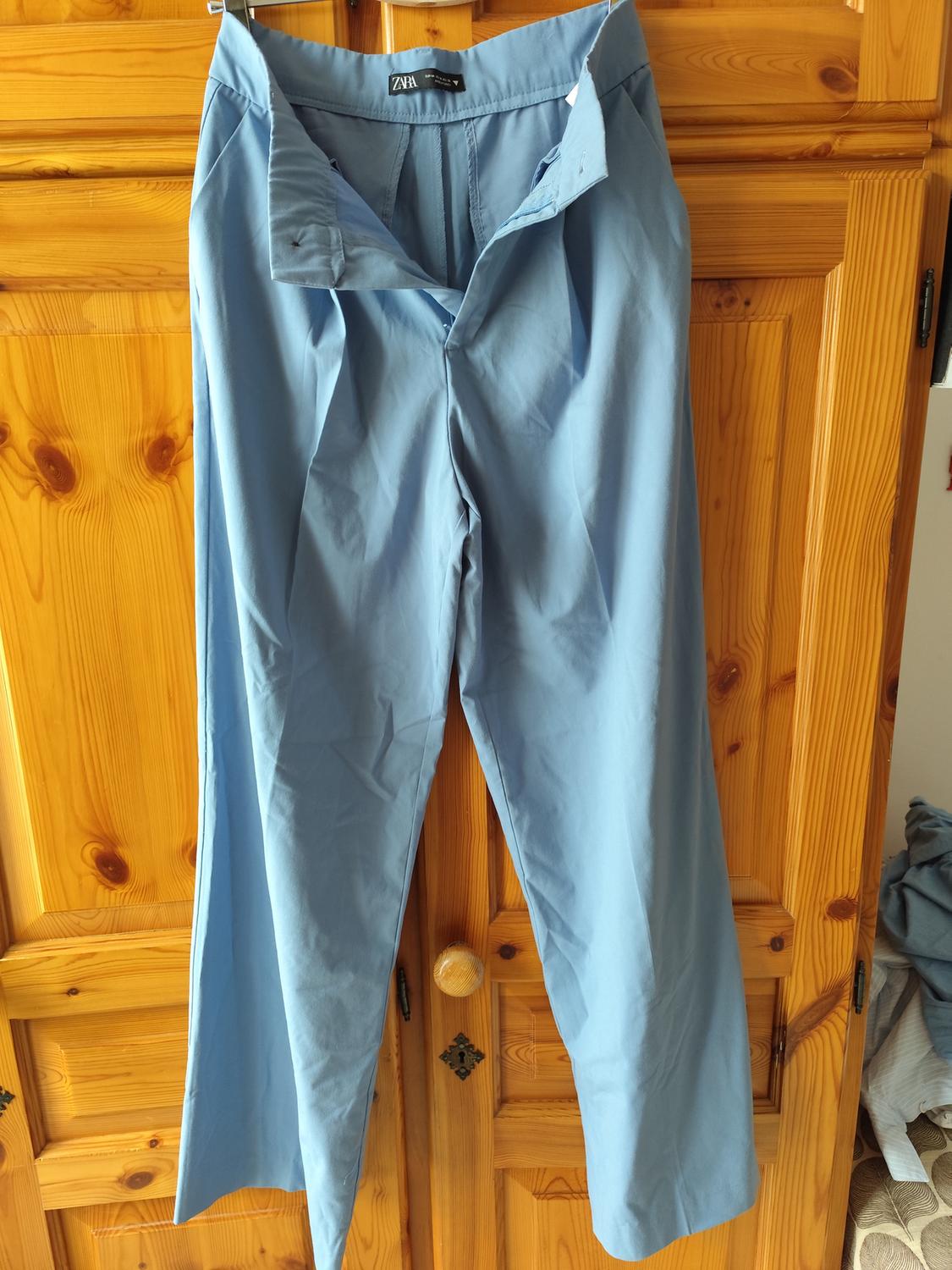 Blue trousers hanging on a wooden door. The label says "Zara". Light wooden background with knot details.