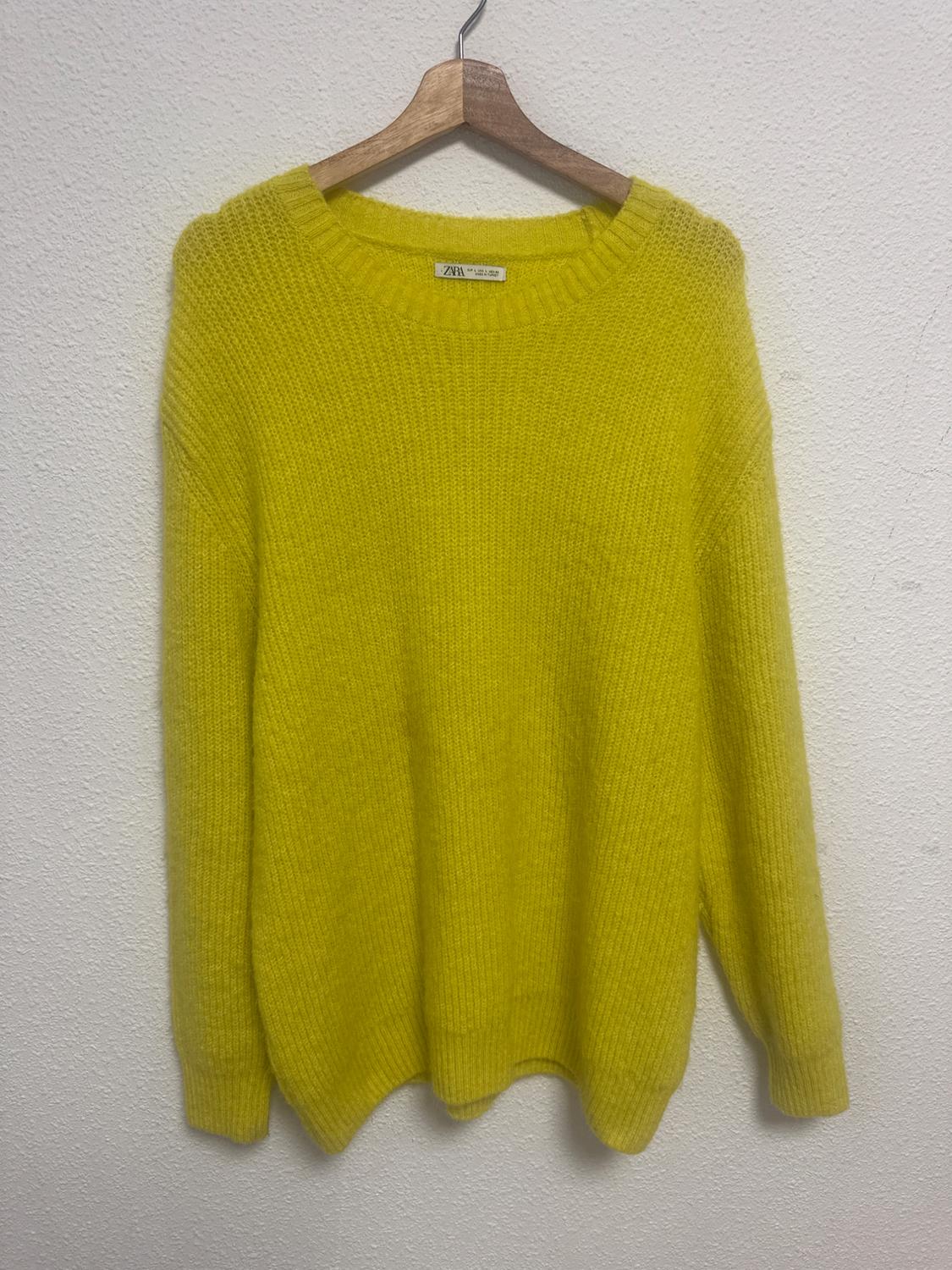 Chunky knit yellow jumper hanging on a wooden hanger against a white wall. Label visible on the collar.