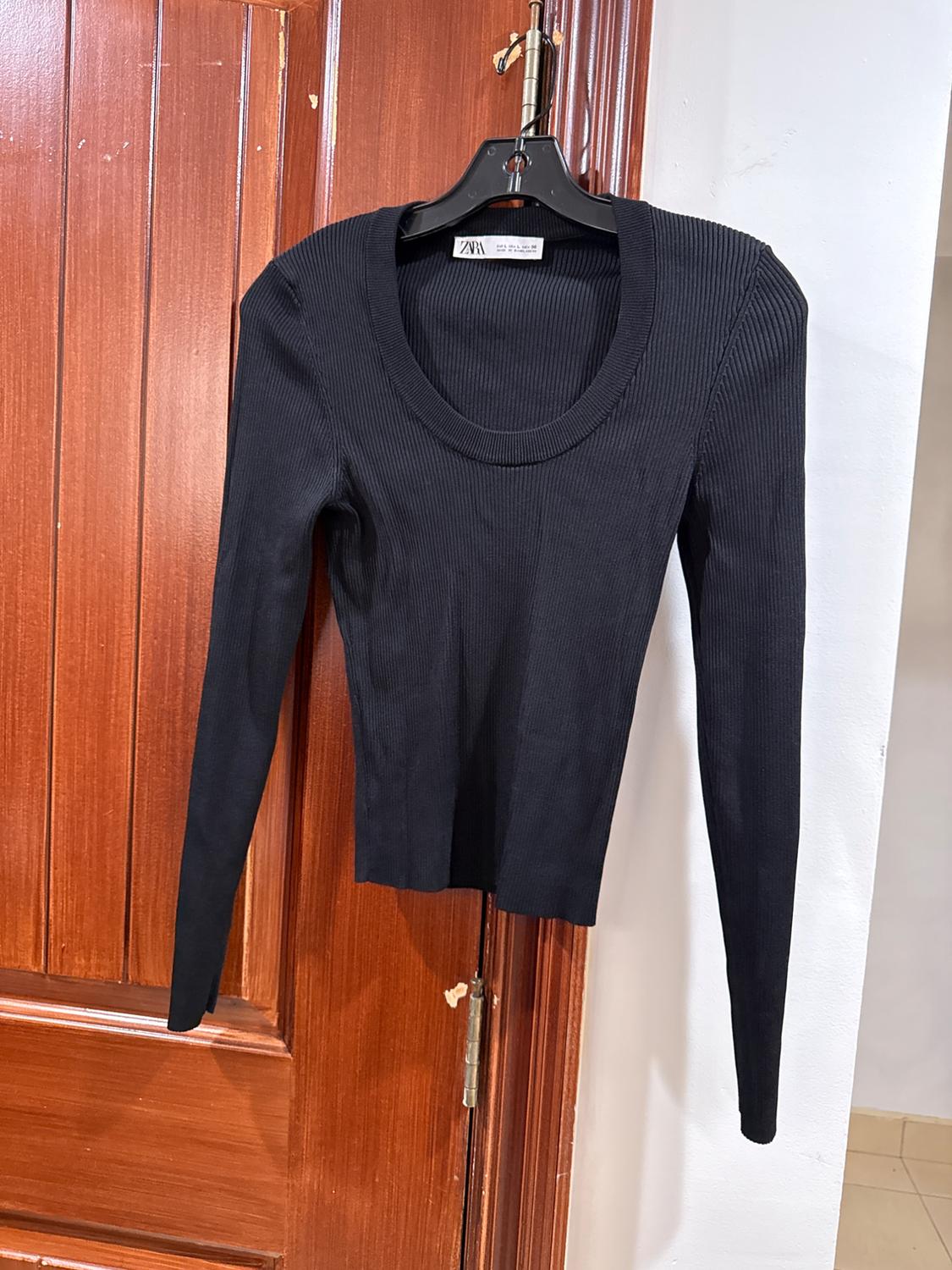 Long-sleeved black sweater hanging on a black hanger over a wooden door. Label visible on the neck.