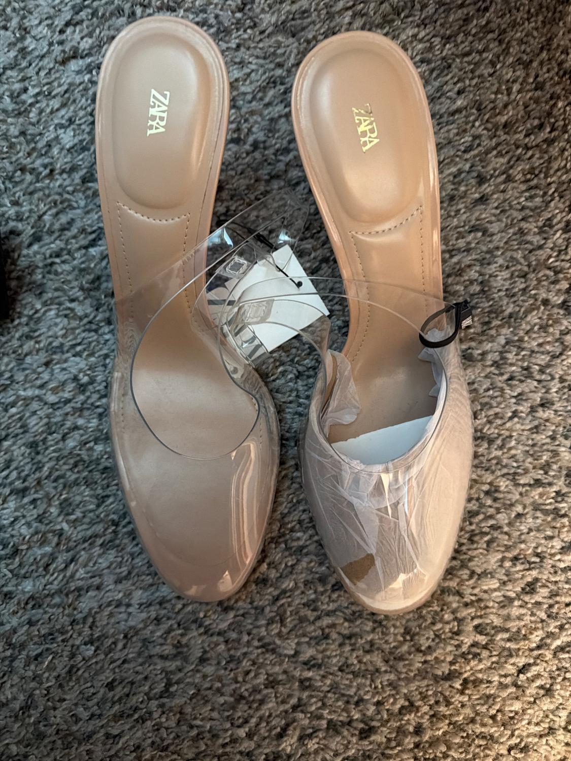 Beige high heels with clear straps on a gray carpet. "ZARA" logo visible on the insole.