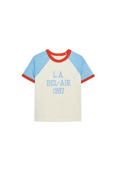 Front view of a white T-shirt with blue sleeves and red trim. Text "L.A. BEL-AIR 1987" on the front.