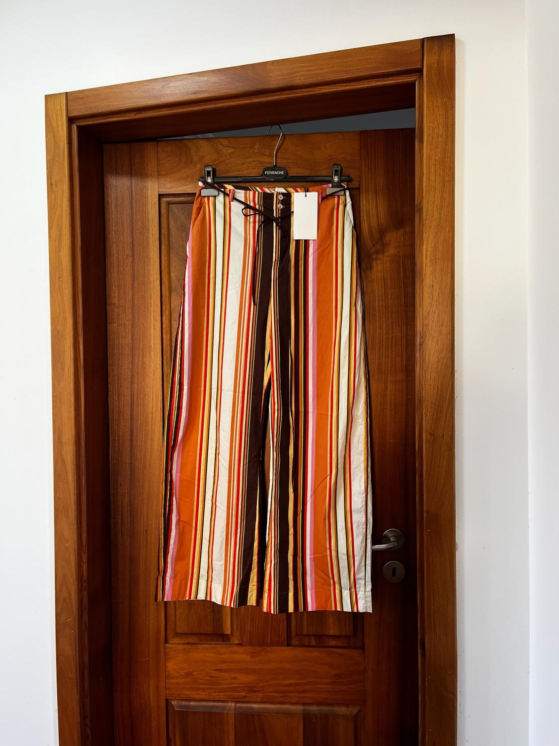 Wide-leg trousers with orange, white and brown vertical stripes hanging on a wooden door. FERRACHE text on the hanger.