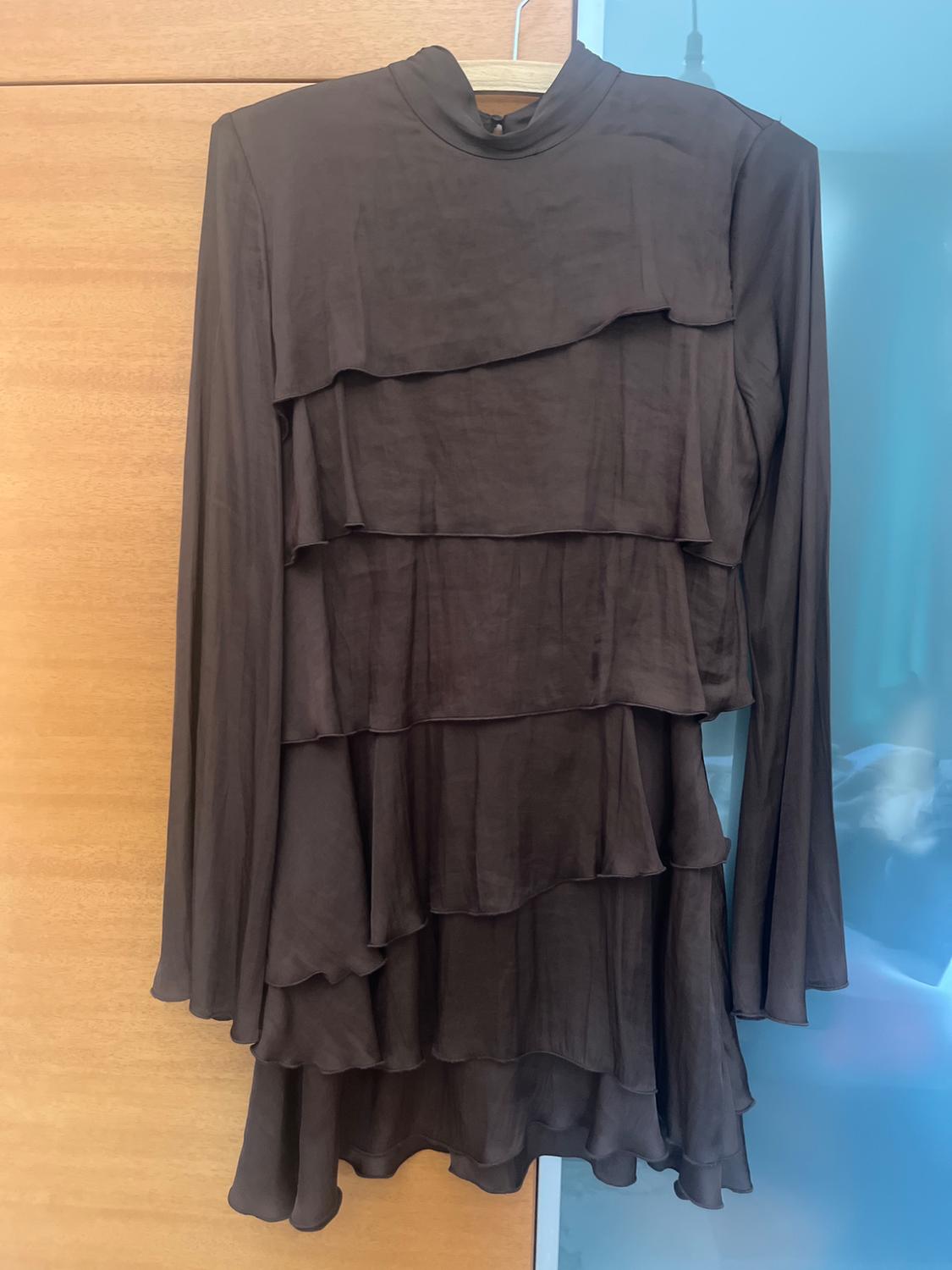 Dark brown, long-sleeved, high-neck mini dress with layered ruffle design, hanging on a hanger against a wooden background.