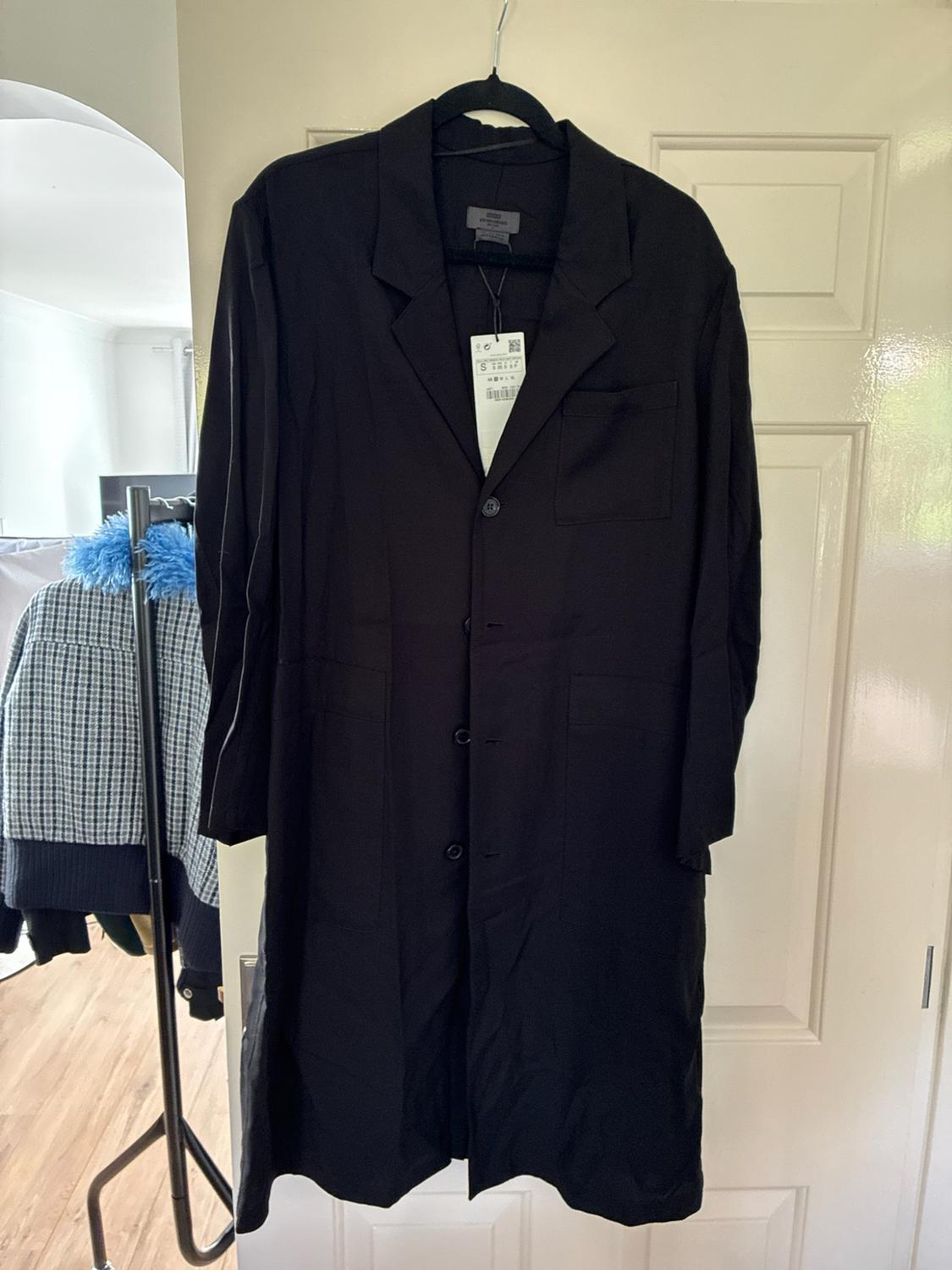Long black coat with lapels and buttons hanging on a white door. In the
background, you can see a clothes rack with clothes in a room.