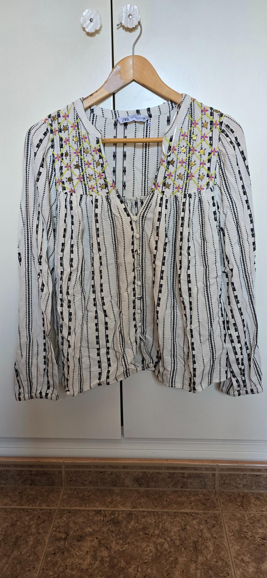 White long-sleeved button-up blouse with black stripes and floral embroidery, hanging on a hanger. Text: ZARA.